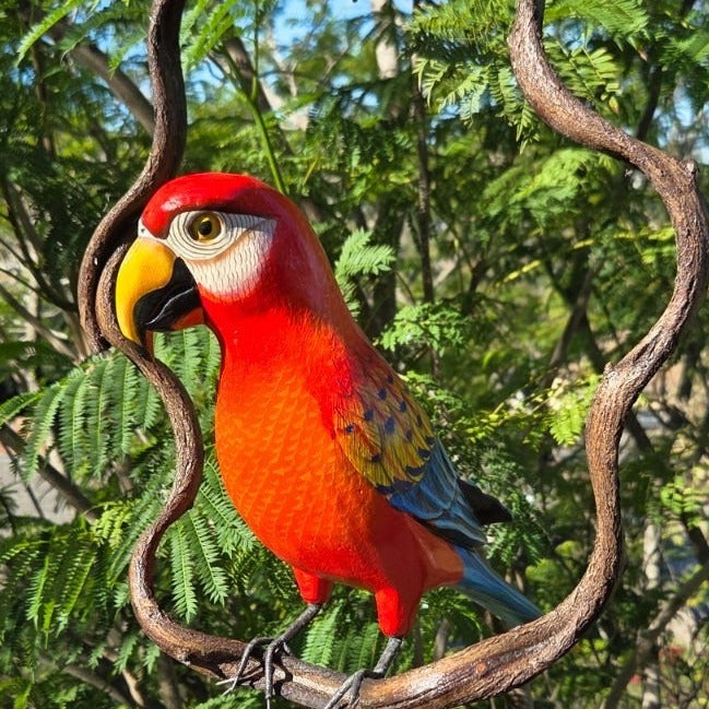 Red wooden parrot perched on a vine branch with green foliage in the background