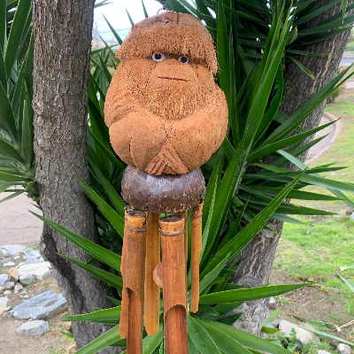 Hand-carved coconut monkey with bamboo wind chimes placed against a natural background with green foliage.
