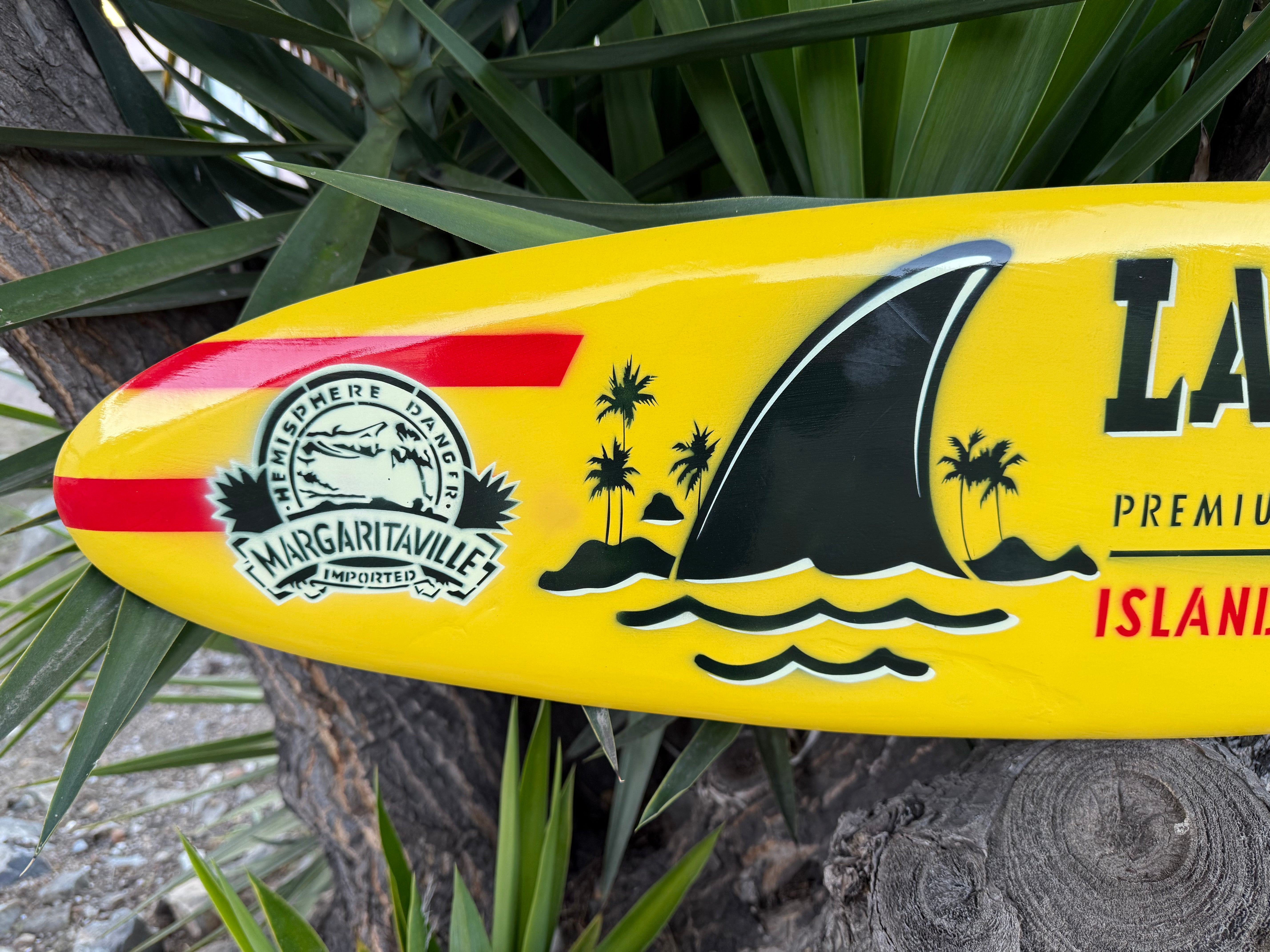 Yellow Margaritaville surfboard with shark design and brand logo against a natural background.