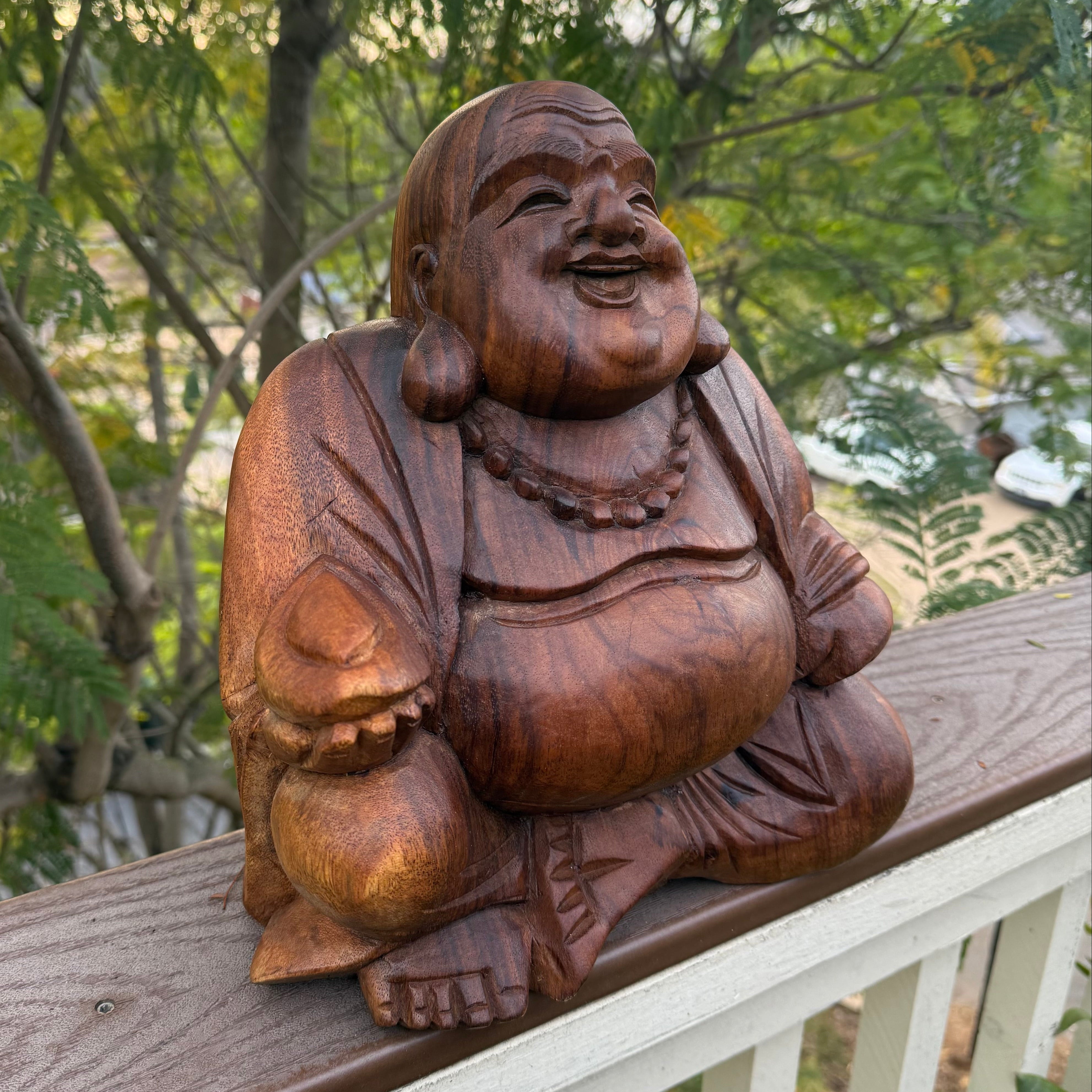 Wooden laughing Buddha statue on a railing with trees in the background