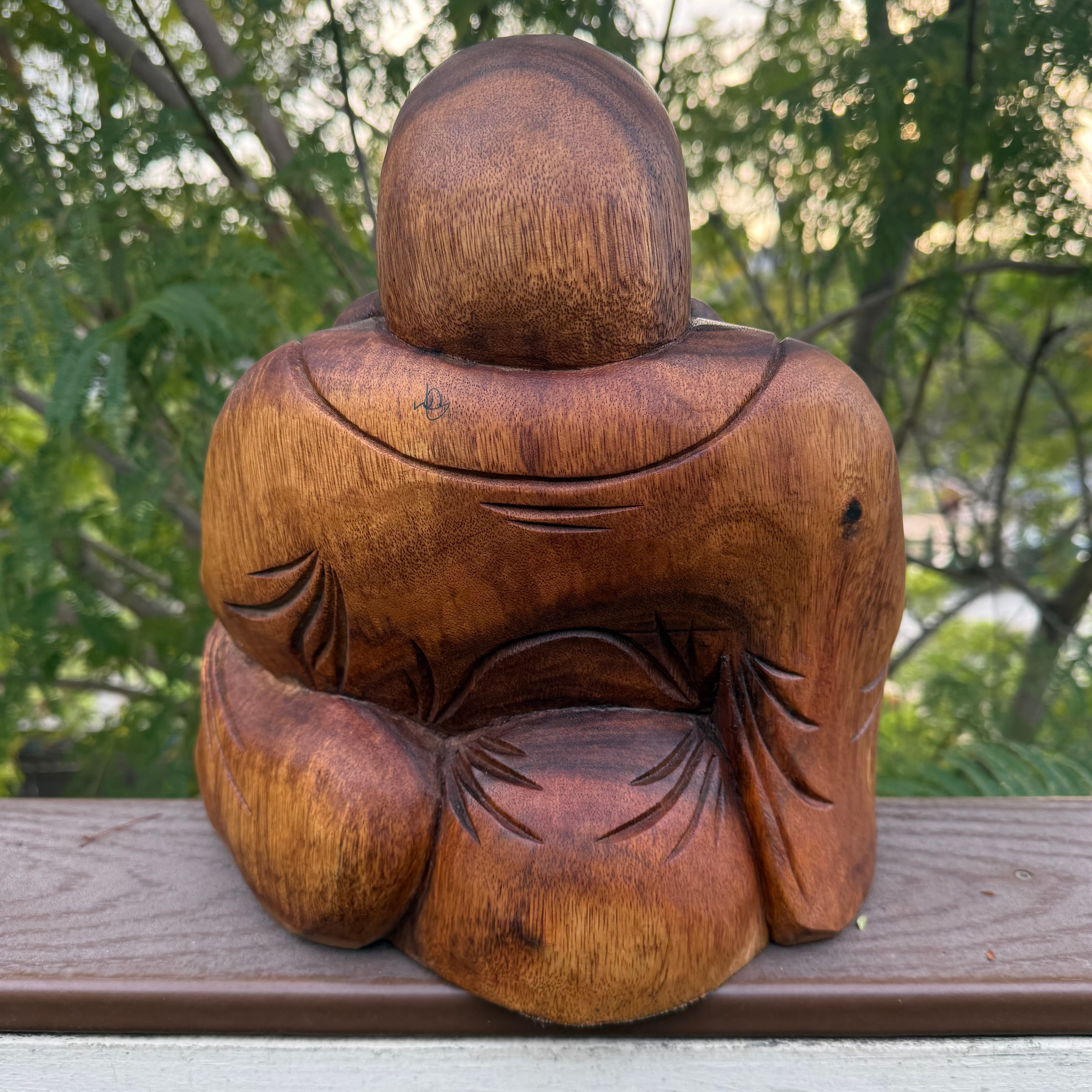 Wooden statue of a seated figure on a wooden surface with greenery in the background