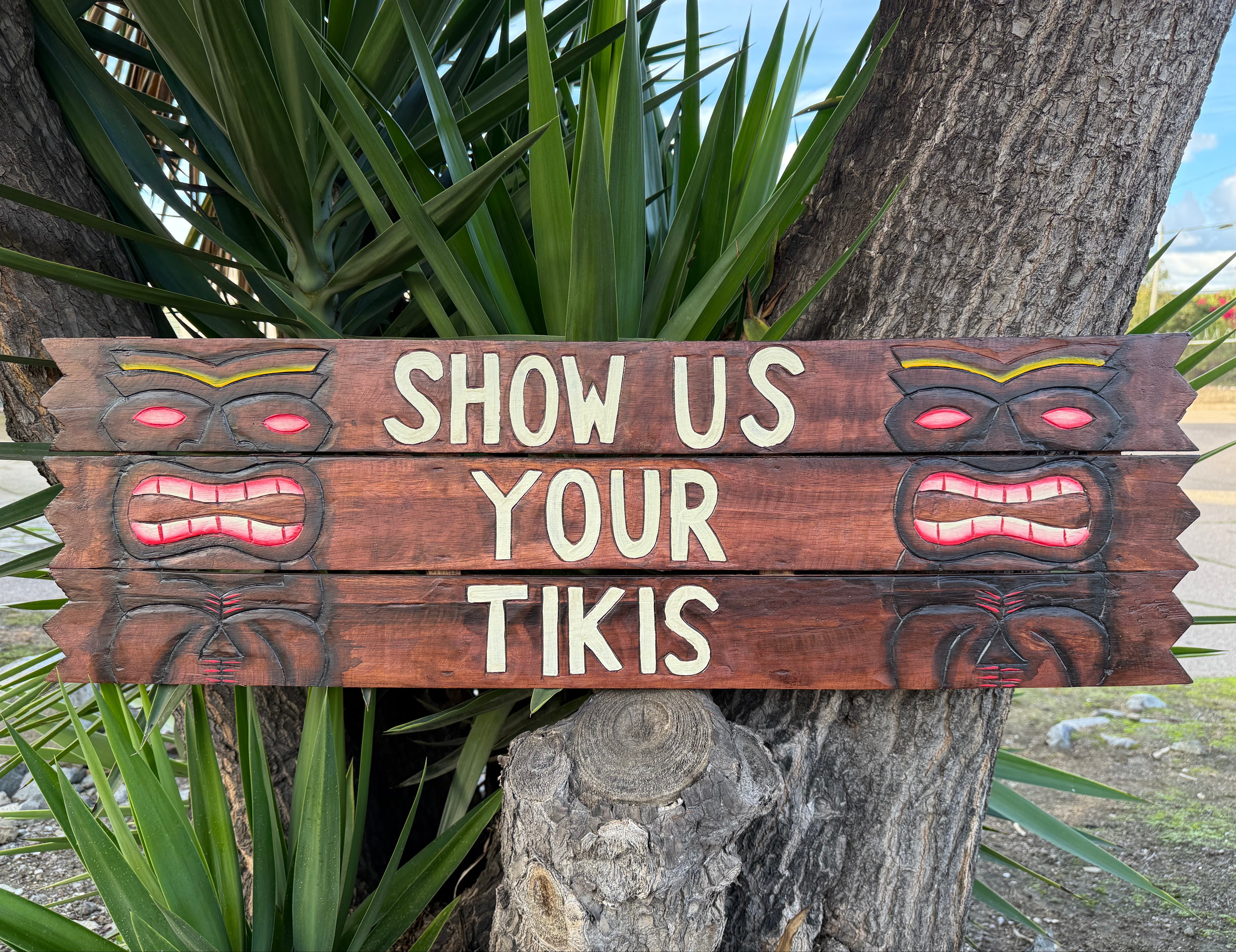 Wooden sign with tiki designs and text 'Show Us Your Tikis' against a natural background.