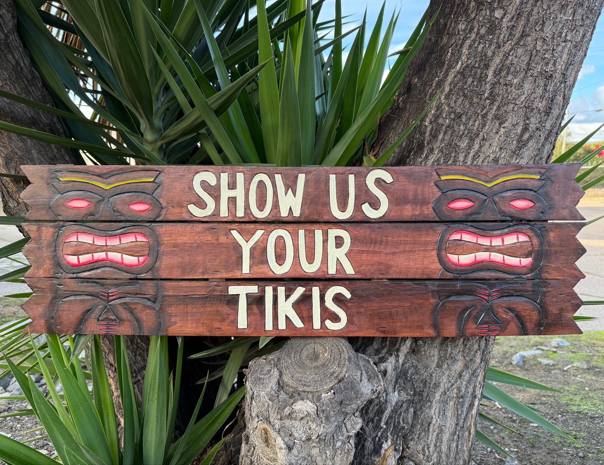 Wooden sign with tiki designs and text 'Show Us Your Tikis' against a natural background.