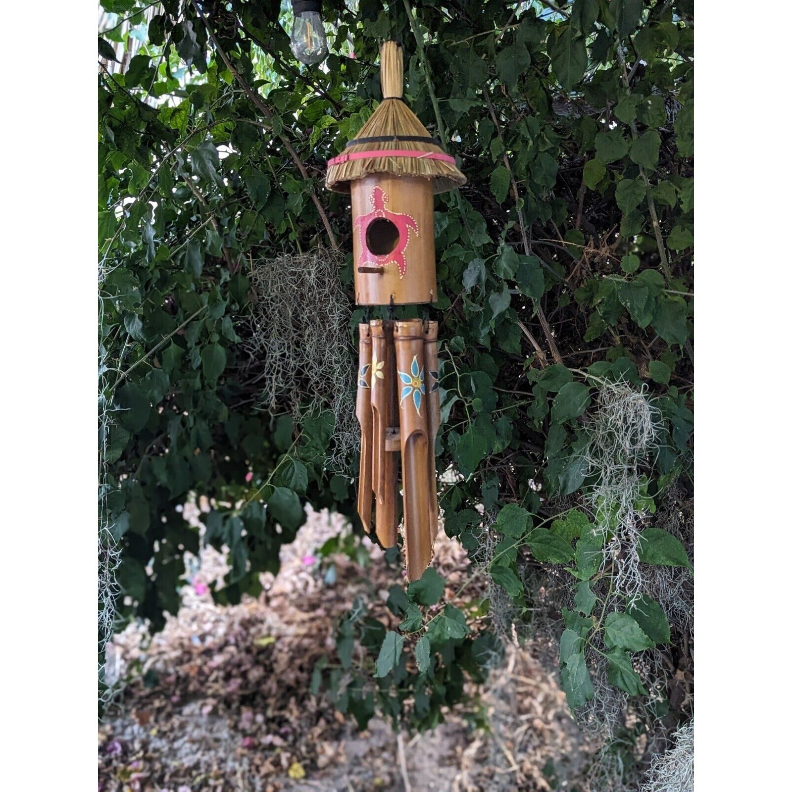 bamboo birdhouse wind chime with straw roof hanging among green foliage