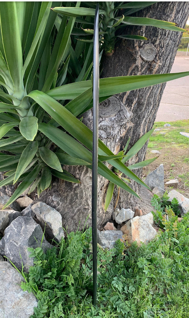 black metal garden stake beside a tree trunk among green plant leaves and rocks