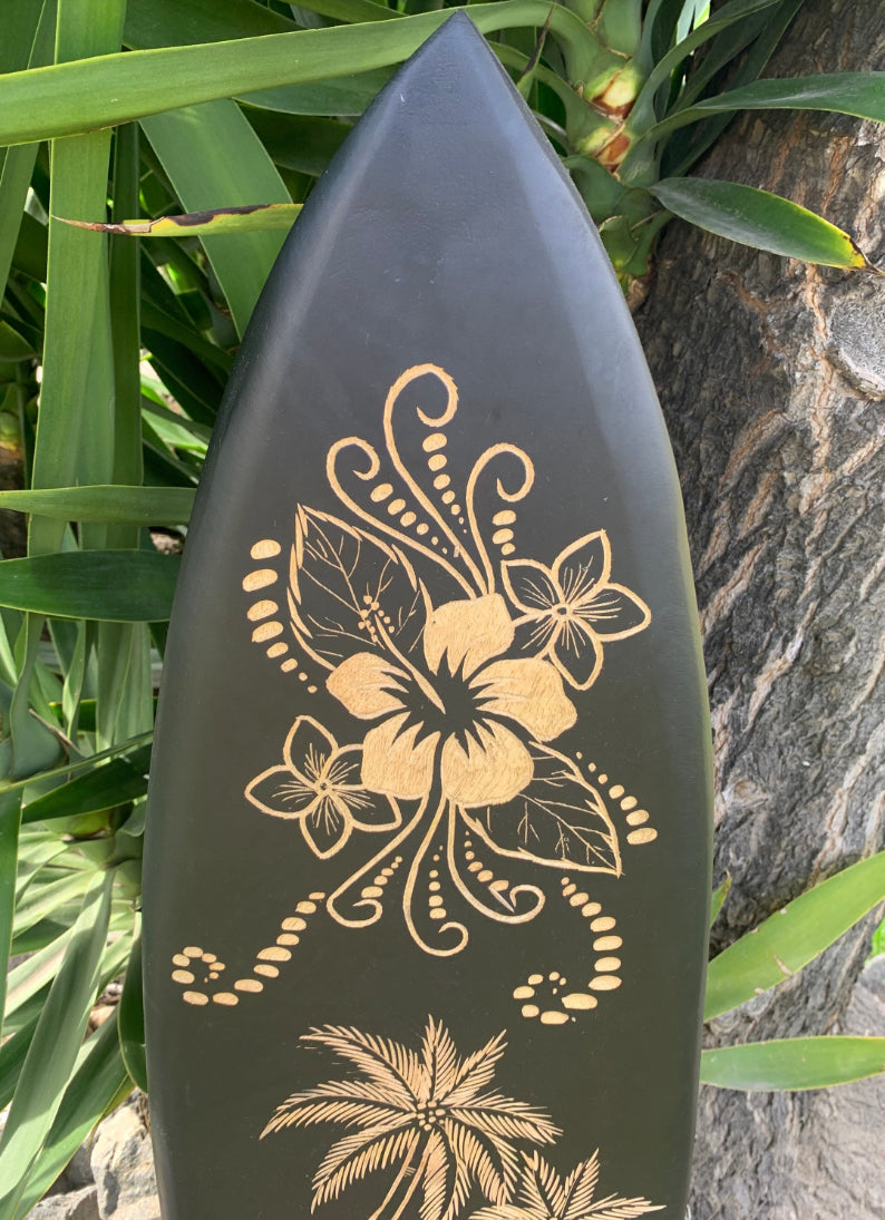 Black surfboard with gold hibiscus and palm-tree artwork leaning against a tree among tropical leaves.