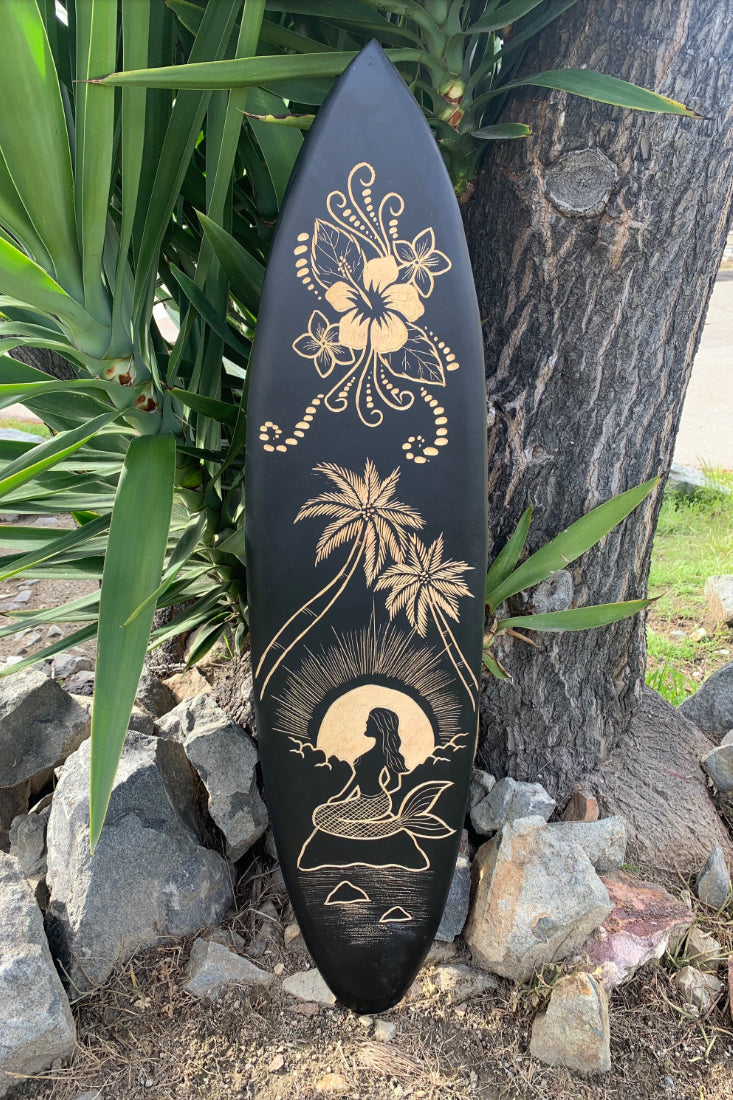 Black surfboard featuring gold tropical hibiscus flowers, palm trees and a mermaid at sunset