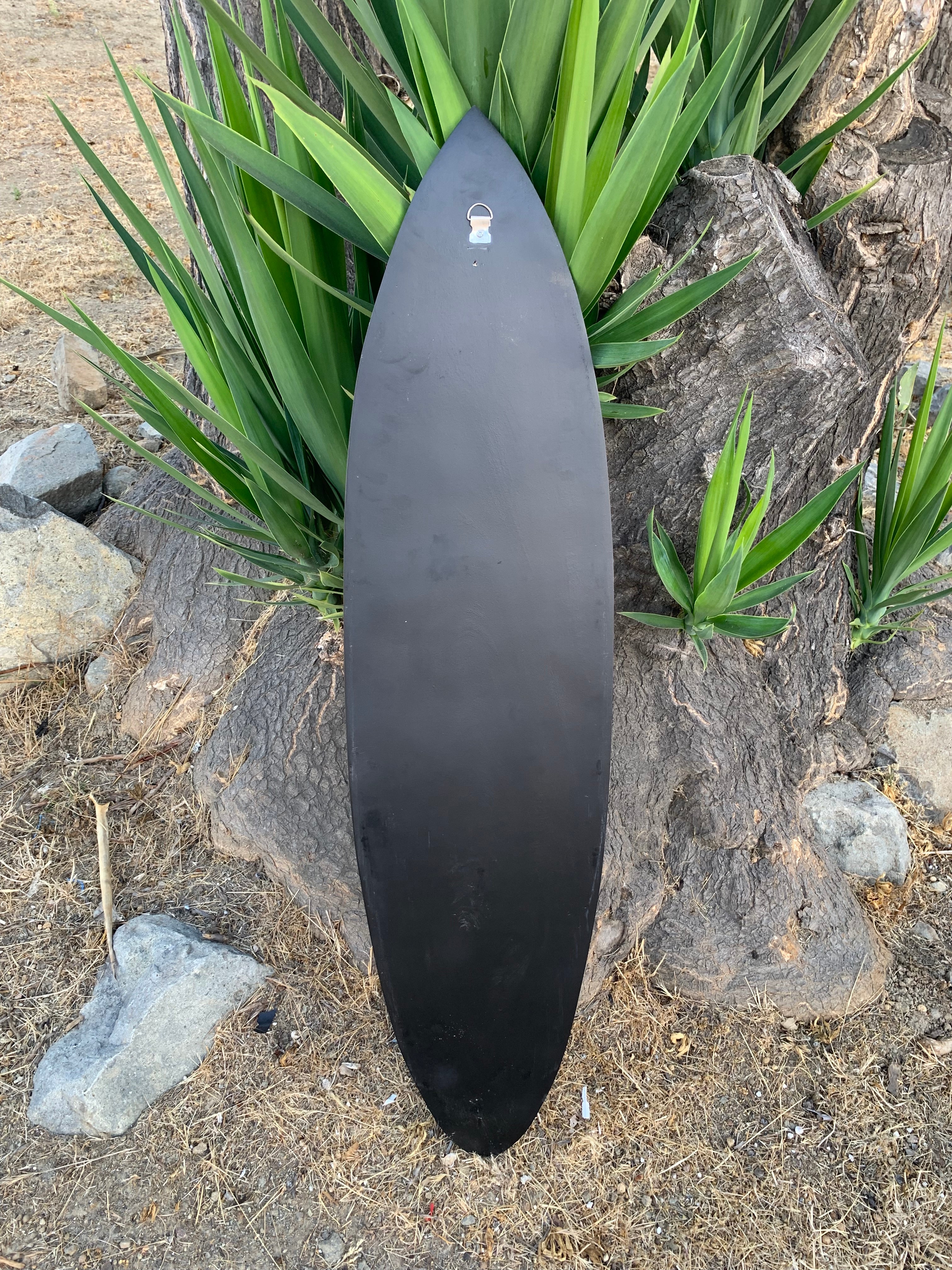 Matte black surfboard propped upright against a tree trunk with green spiky plants around.
