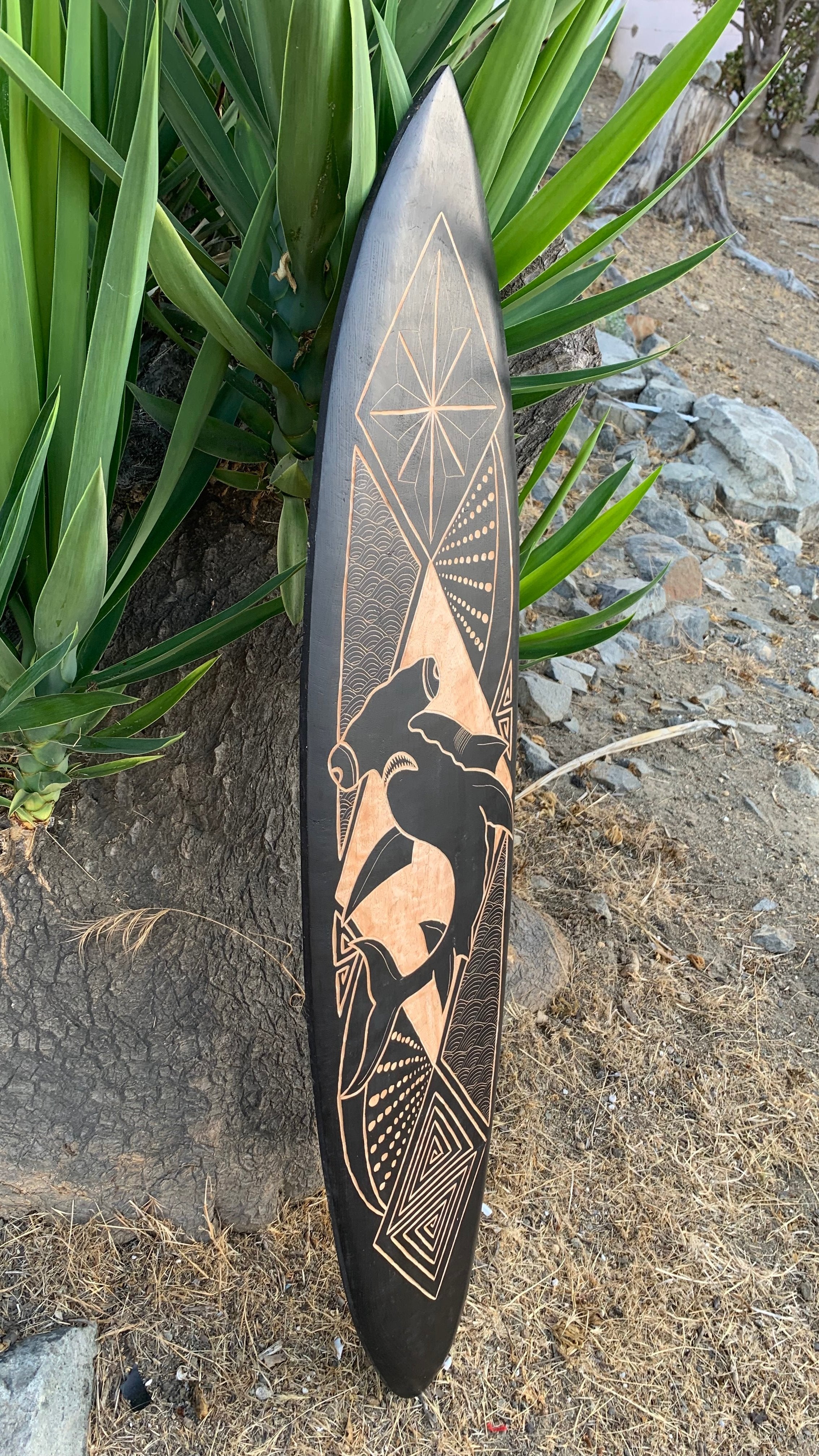 Black surfboard with copper tribal etching and geometric patterns leaning by a plant