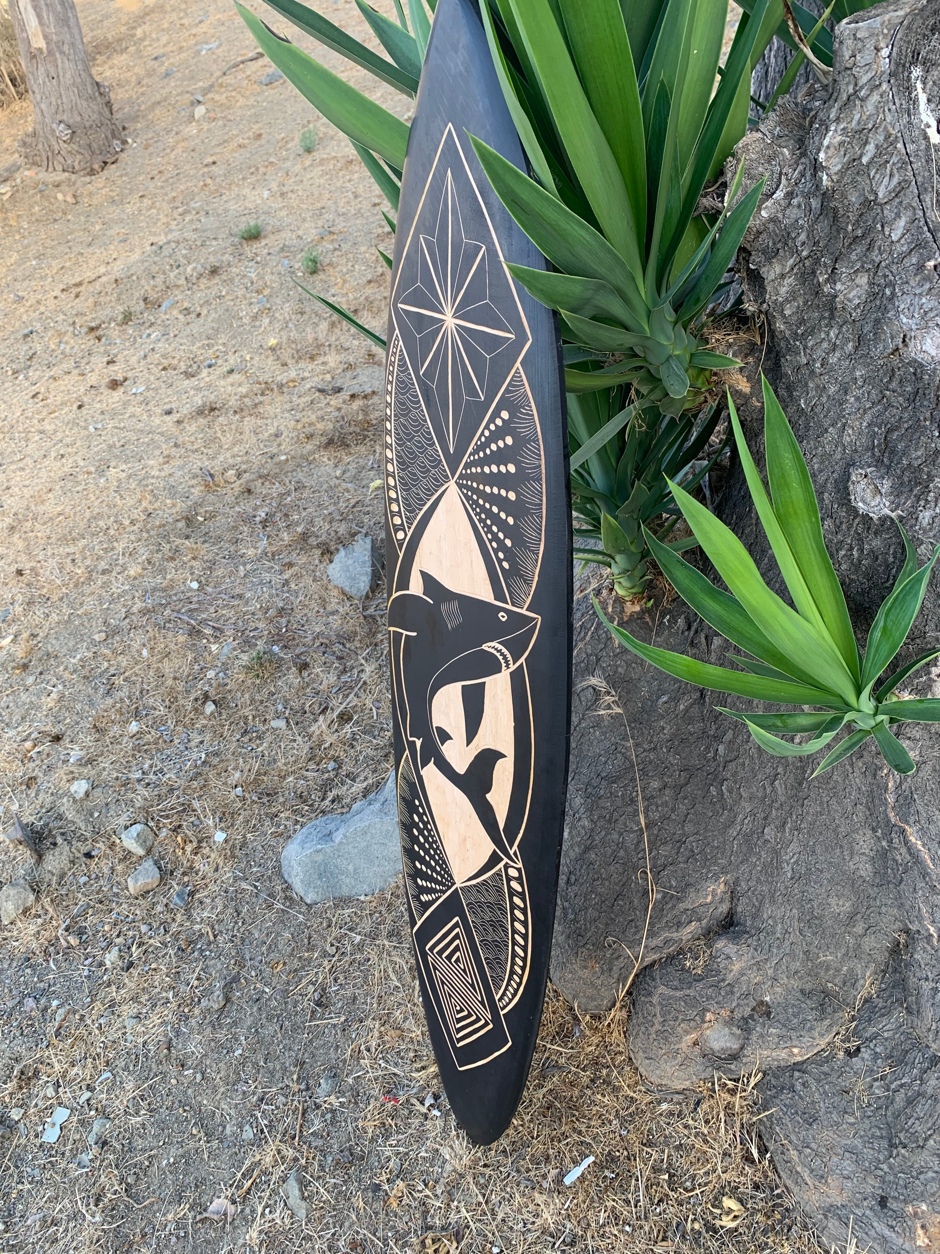 Decorative black surfboard with tan geometric patterns leaning against a tree among green plants