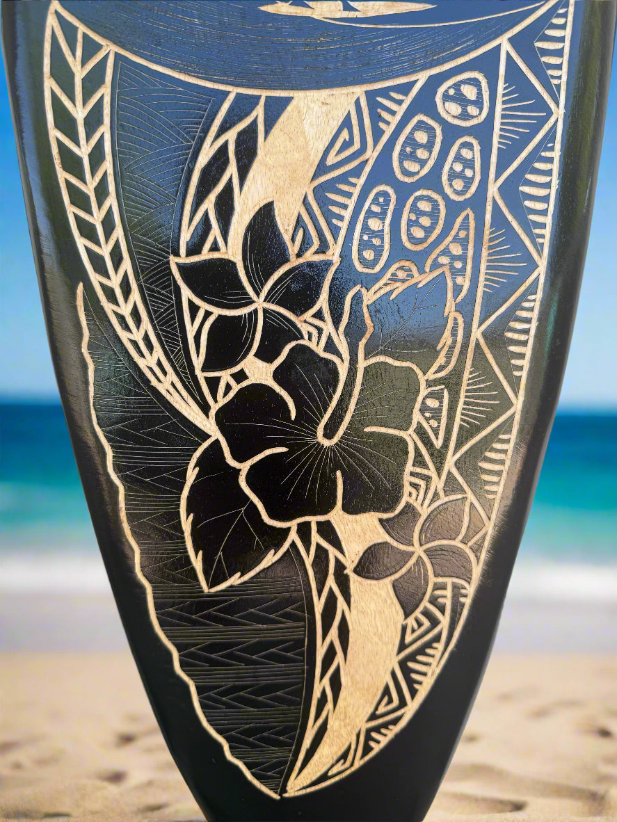 Close-up of a black surfboard with gold hibiscus and leaf engraving on a sunny beach