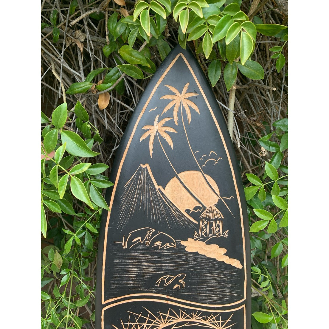 Black surfboard with copper-toned island scene: palm trees, a hut, volcano, and waves, framed by green leaves.