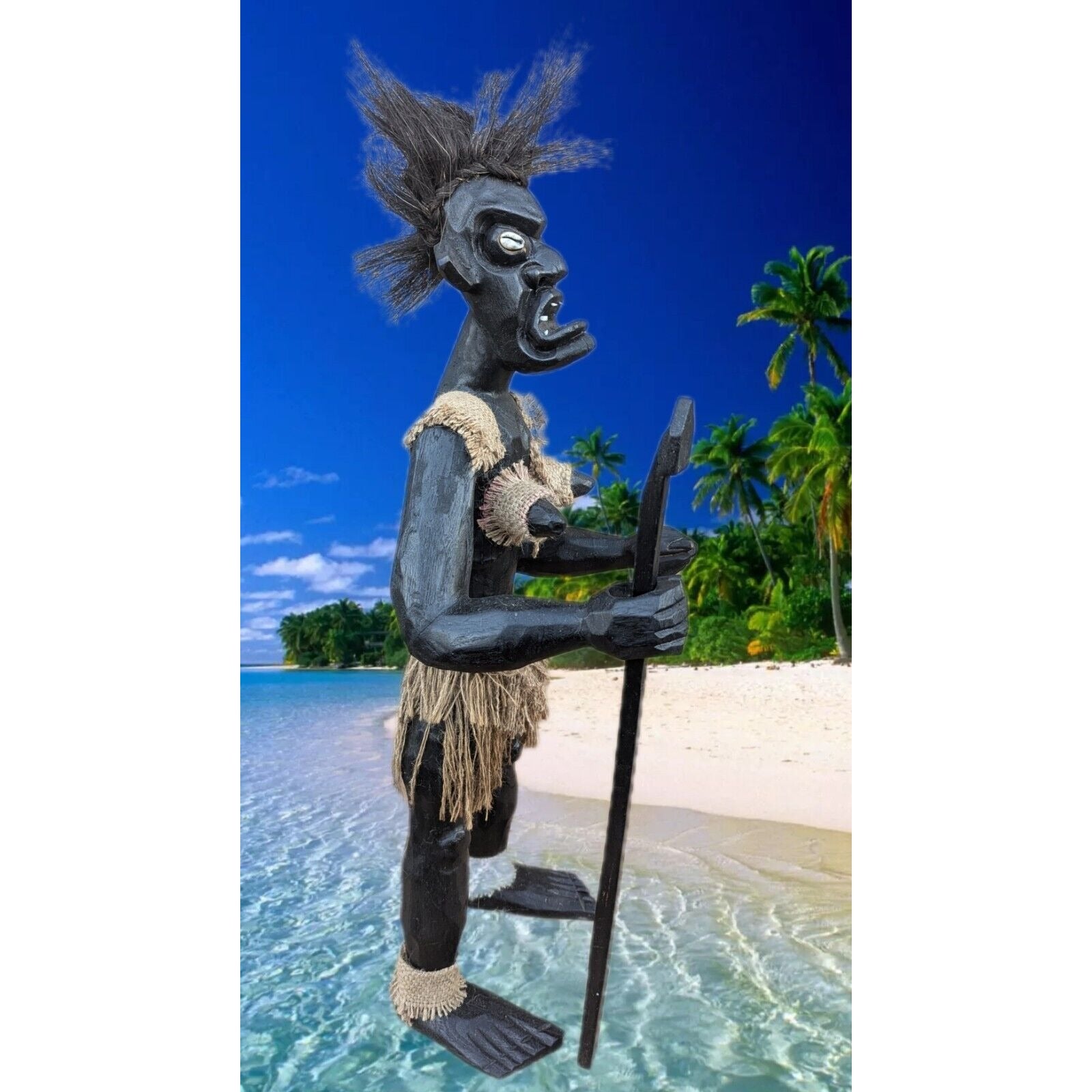Black tiki statue wearing burlap skirt on a tropical beach, holding a spear with palm trees nearby.