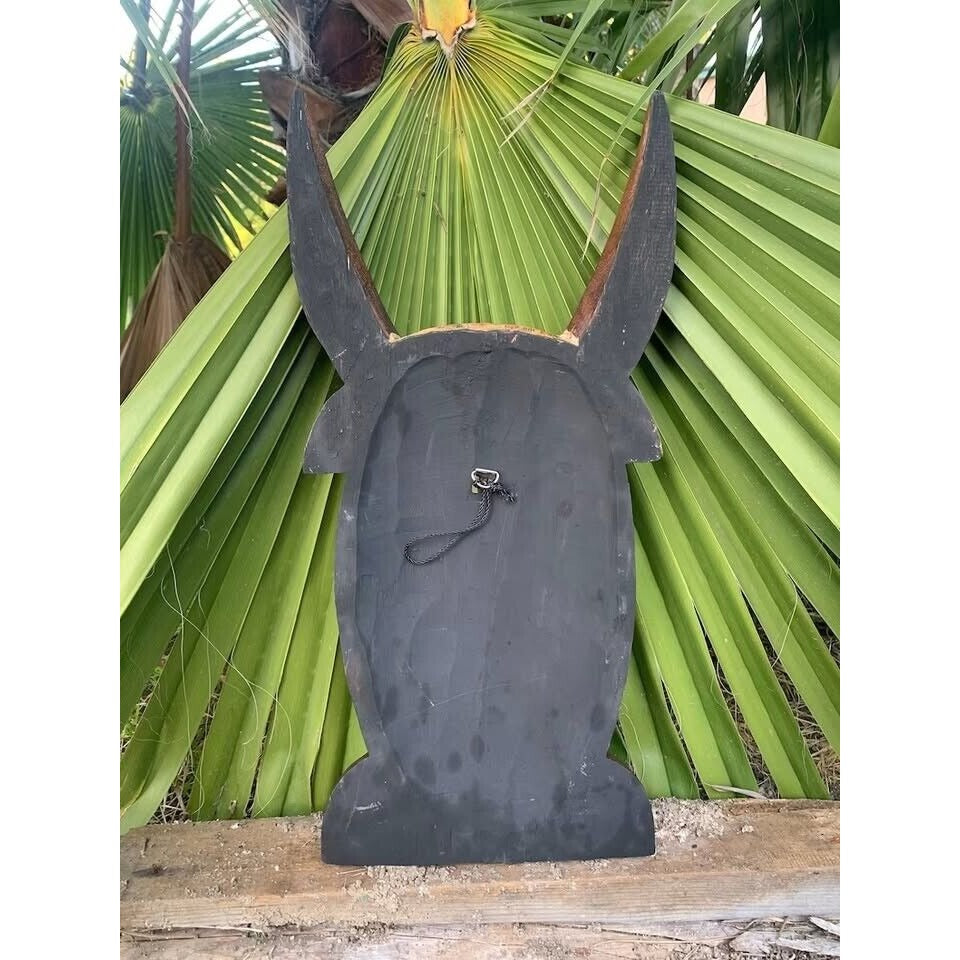 Black wooden donkey head silhouette wall sculpture with long ears against green palm leaves.