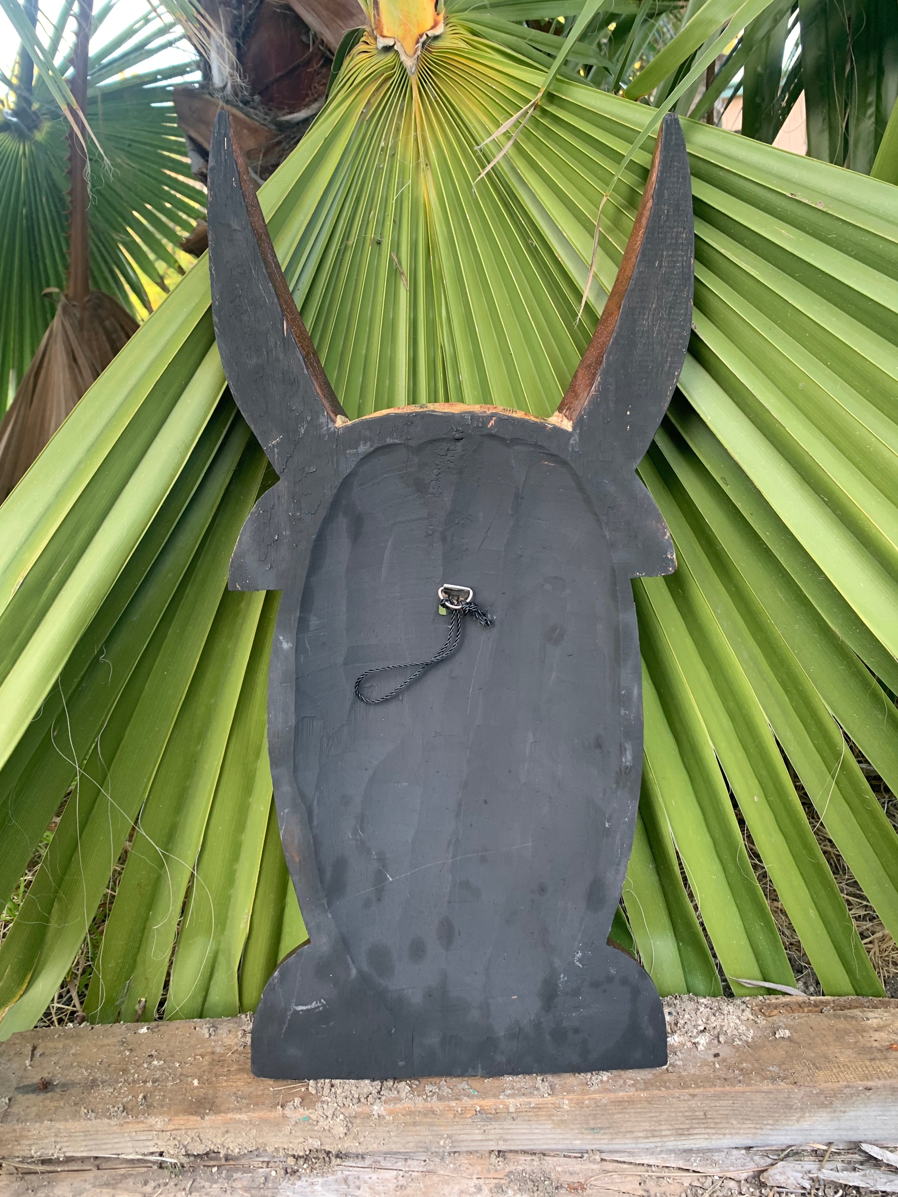Black wooden rabbit silhouette decorative cutout with long ears and hanging loop against green palm leaves.