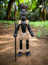 Black wooden tribal figure with spear, burlap shoulder coverings and skirt in a forest setting.