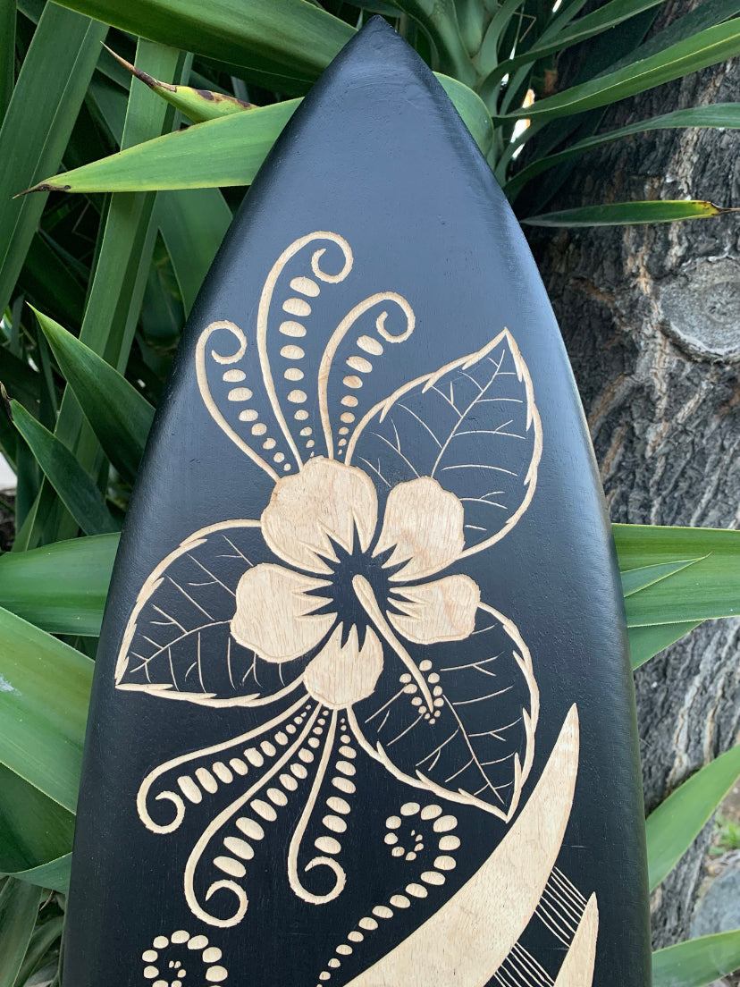 Blue-black surfboard featuring a beige hibiscus carving and tropical leaves.