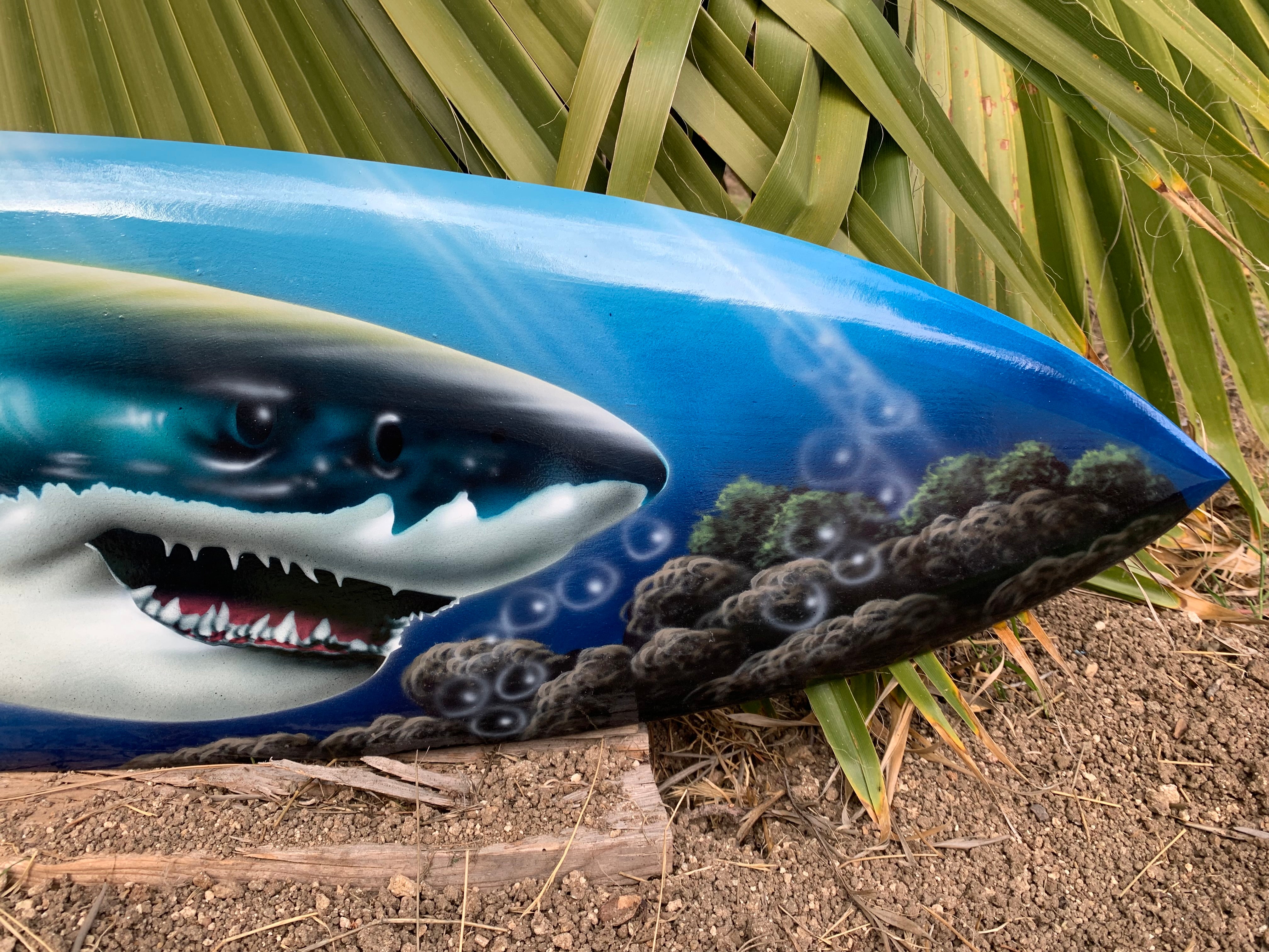 Blue surfboard with a shark illustration amid bubbles and rocks