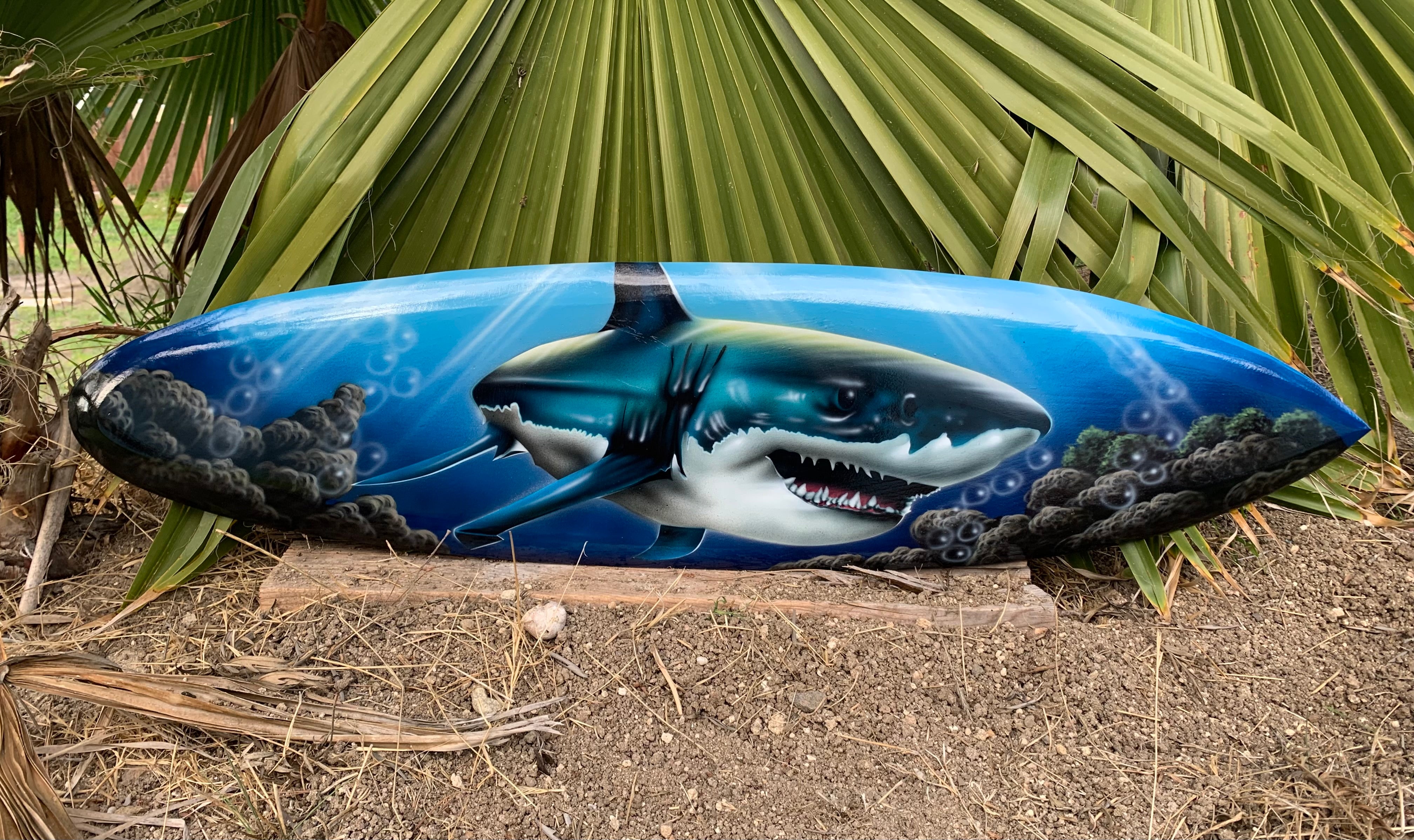 Blue surfboard with a painted shark design resting among palm leaves.