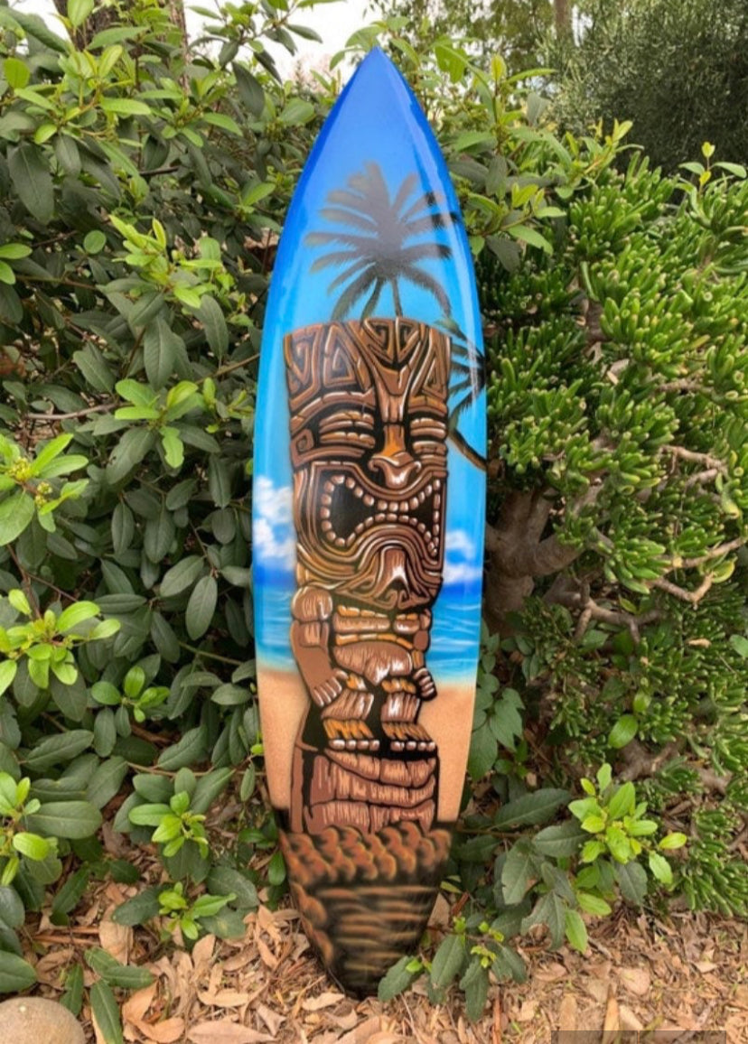 Blue surfboard featuring a carved tiki figure on a tropical beach scene