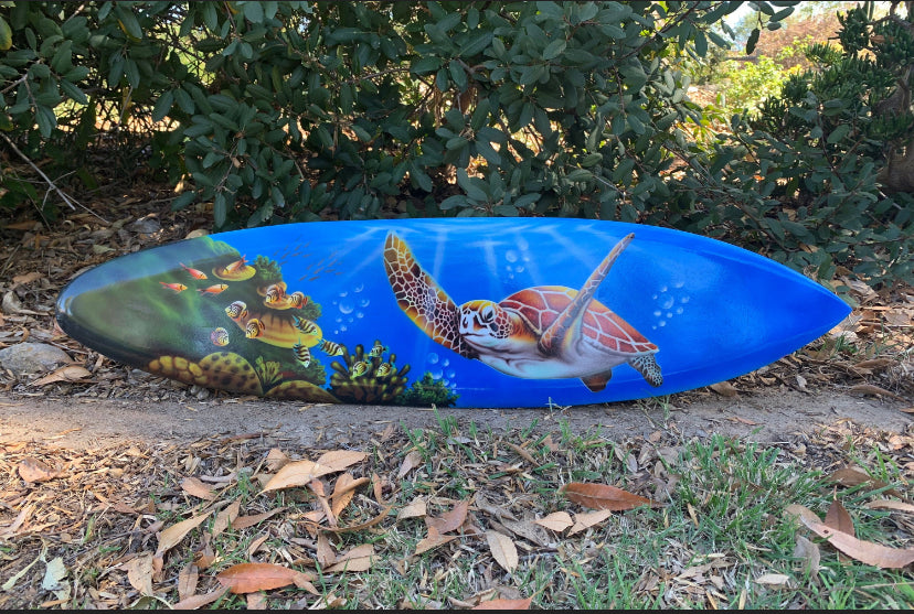 Blue surfboard featuring a sea turtle and coral reef artwork lying on the ground beside leaves