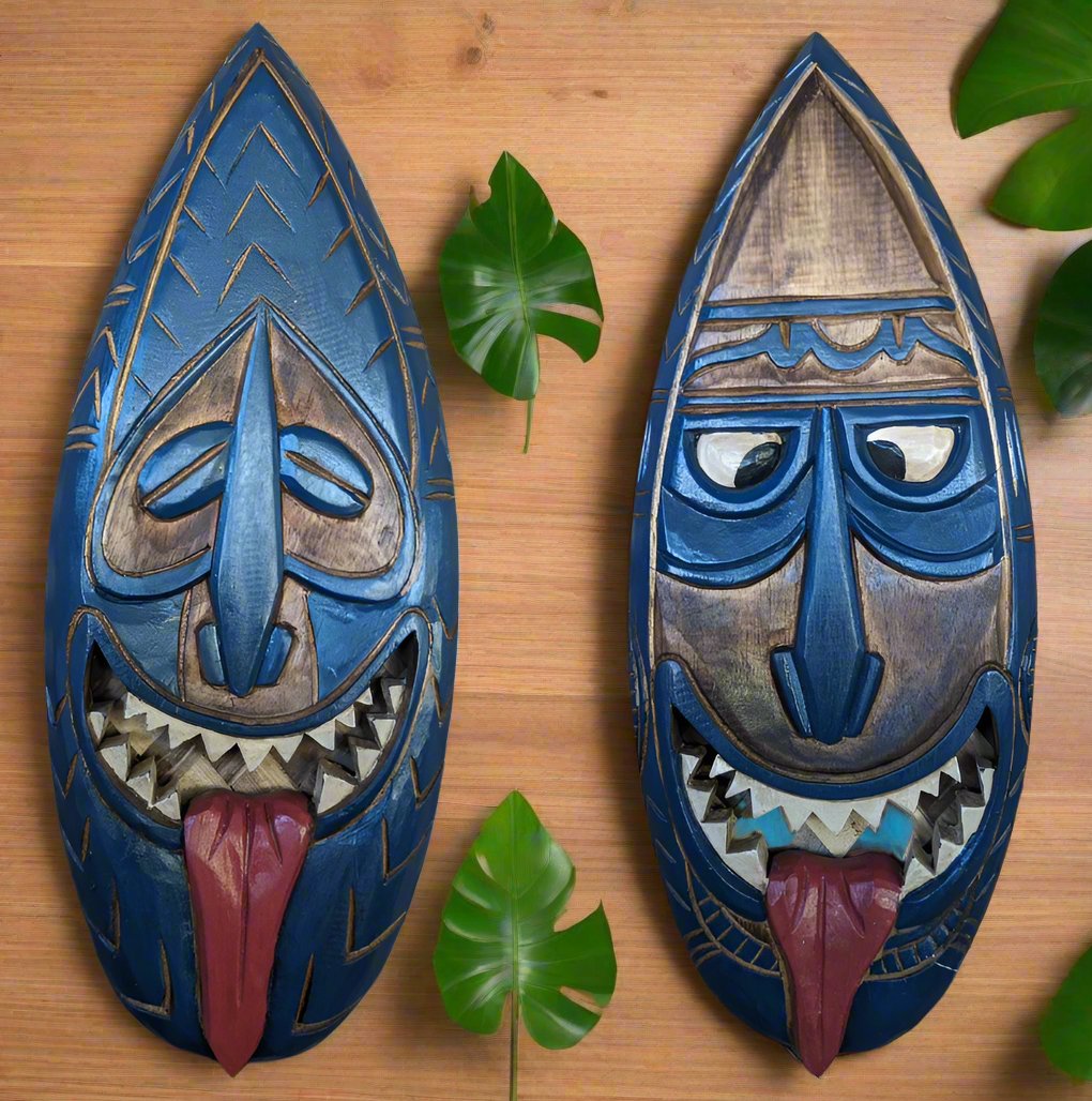 Pair of blue wooden tiki masks with exaggerated features and tongues sticking out on a wooden surface.