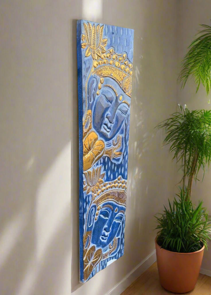 Blue-toned Buddha-inspired wall panel with golden faces and lotus motifs beside a potted plant.