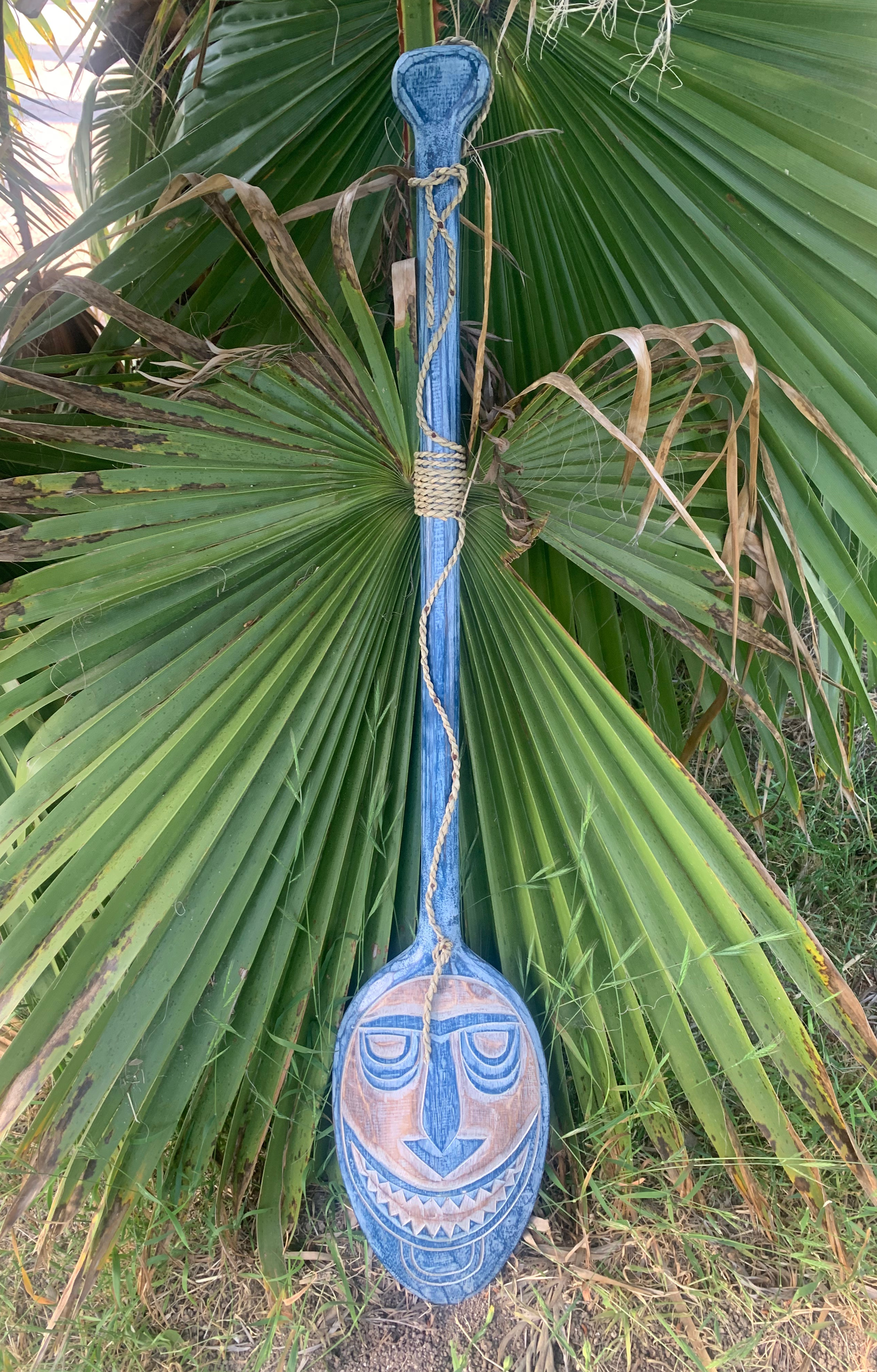Blue wooden oar with a carved face mask hanging among palm leaves