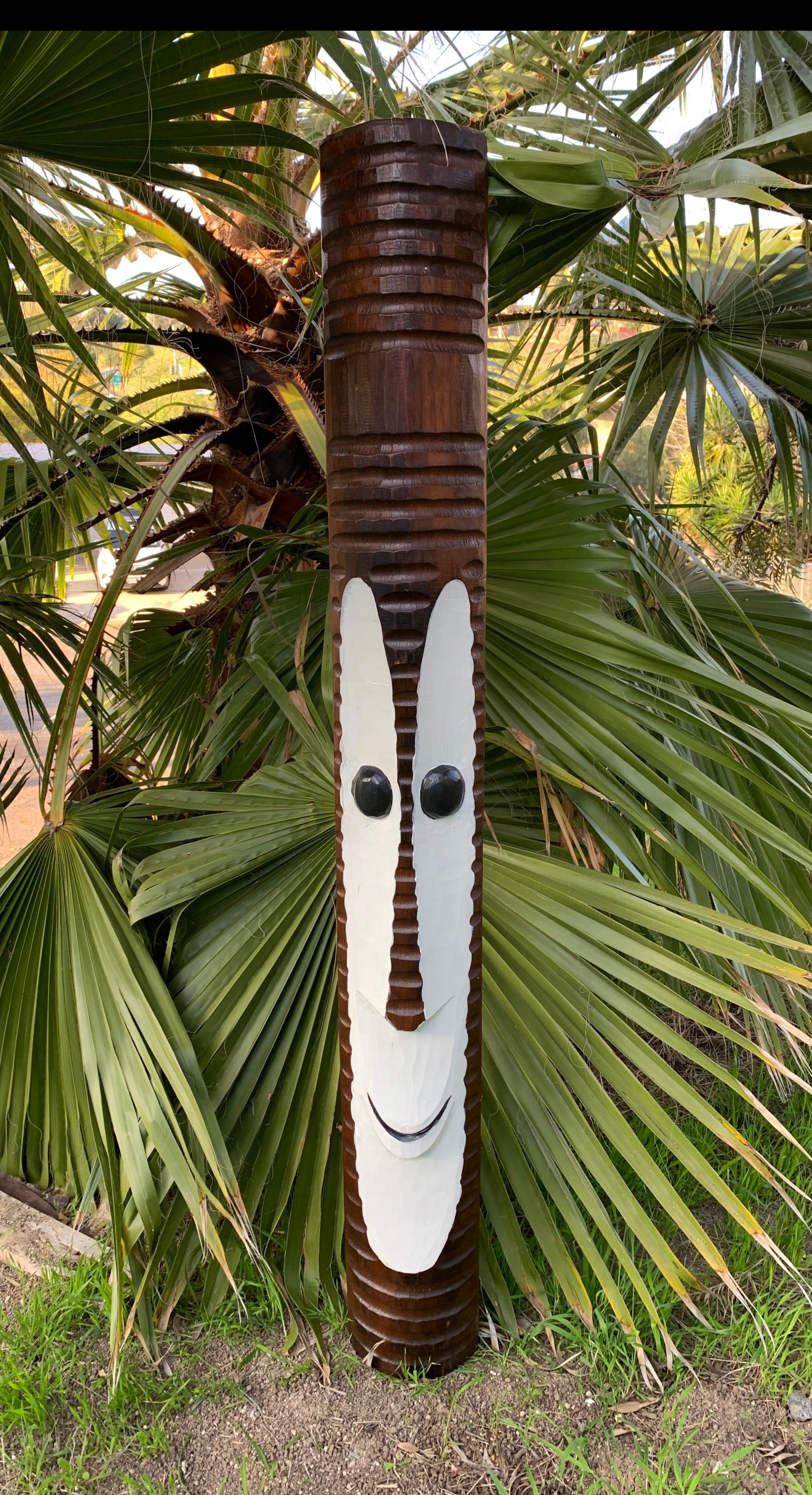 Brown carved totem pole with white face and black eyes among palm fronds in a garden.