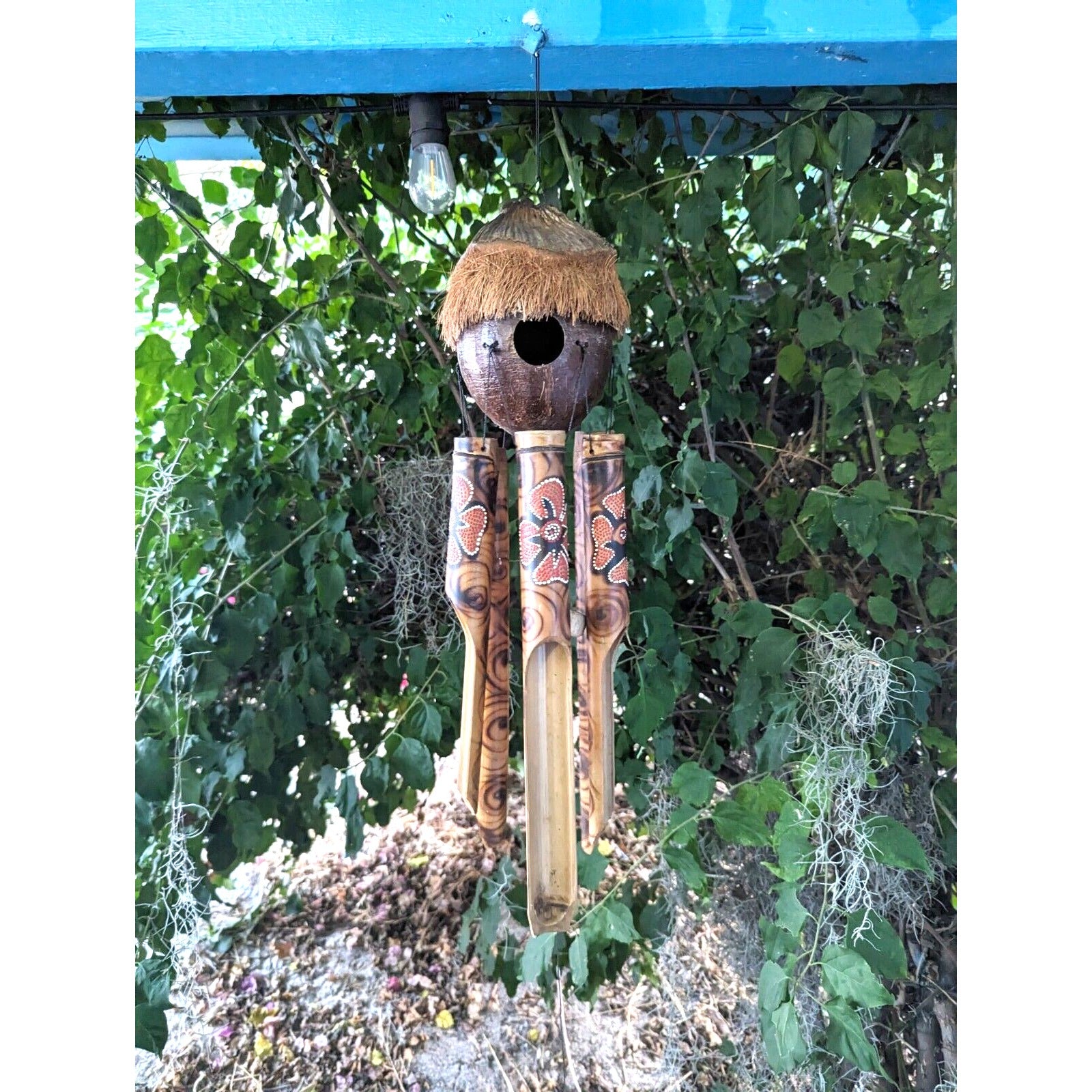 Outdoor wind chime with four carved bamboo tubes and a coconut-shell top with straw fringe, hanging among green leaves