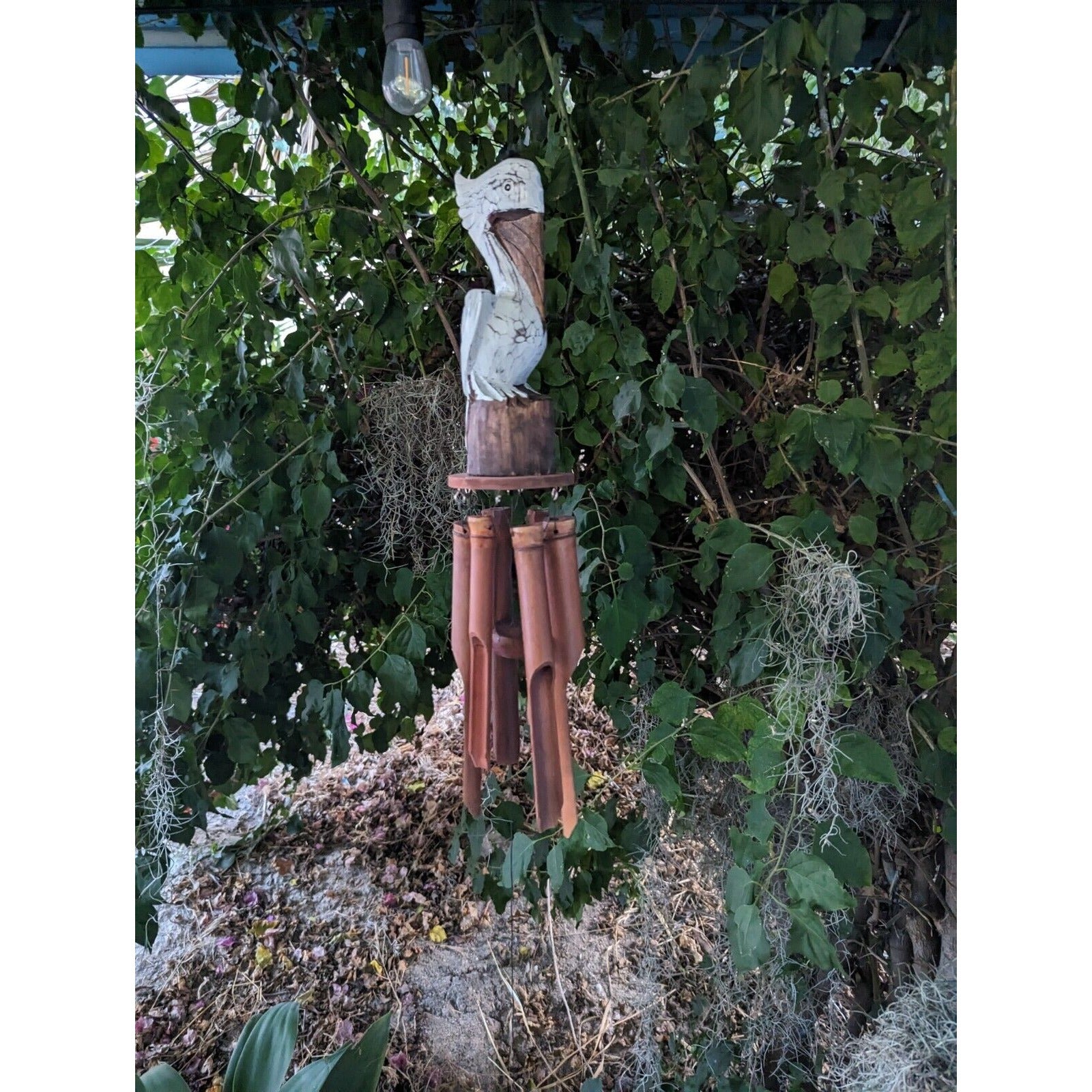 carved bird statue on wind chime with terracotta tubes hanging from a leafy tree