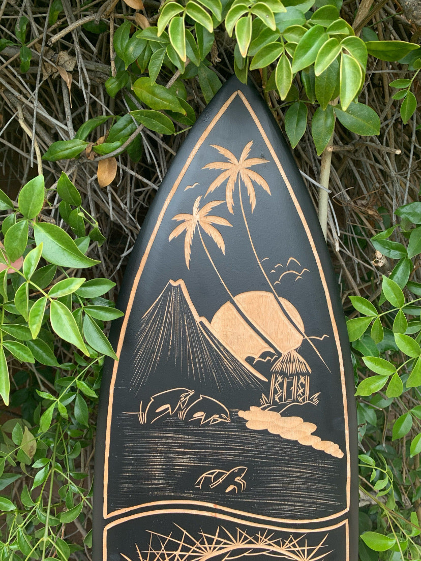 Carved surfboard with palm trees, hut, and volcano beach scene on a dark background among green foliage.