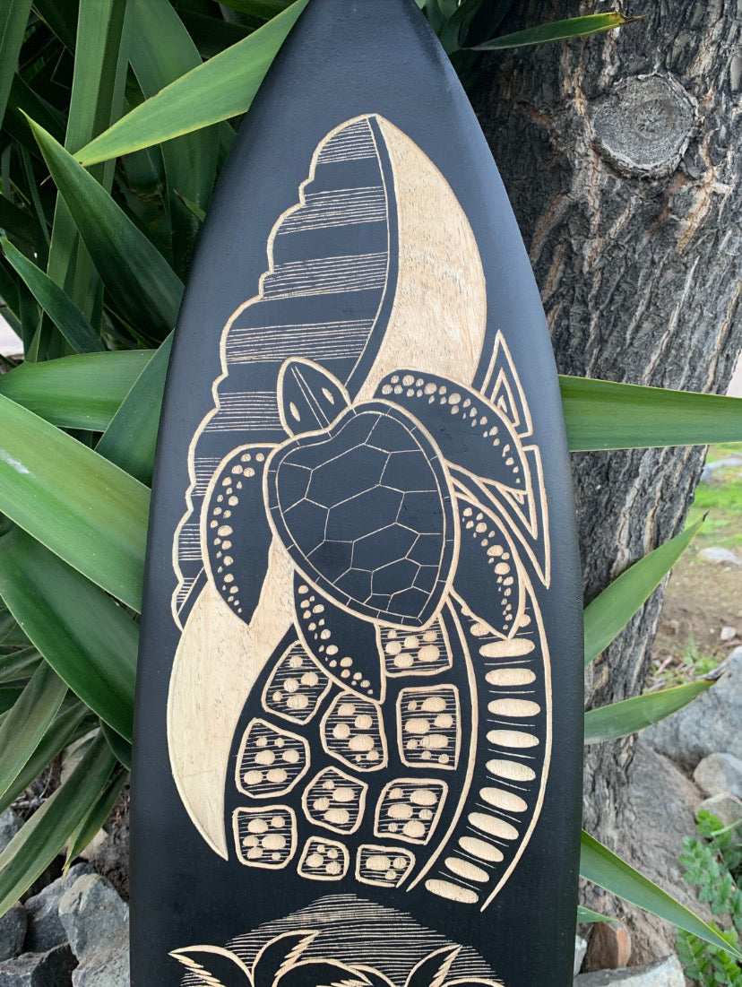 Dark surfboard featuring a beige carved sea turtle design, leaning by a tree with tropical plants.