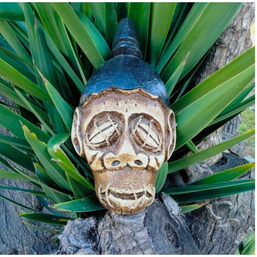 Carved tiki mask attached to a tree trunk among green leaves.