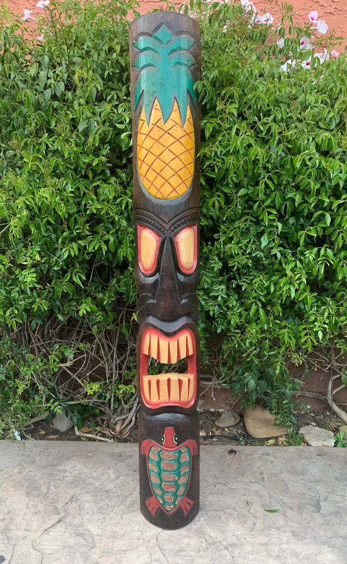Carved tiki totem pole with pineapple crown, red facial features, and a turtle motif at the base.