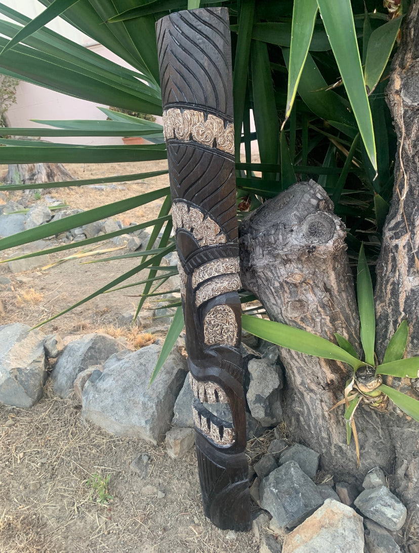 Carved black and tan wooden totem pole with tribal patterns beside rocks and plants
