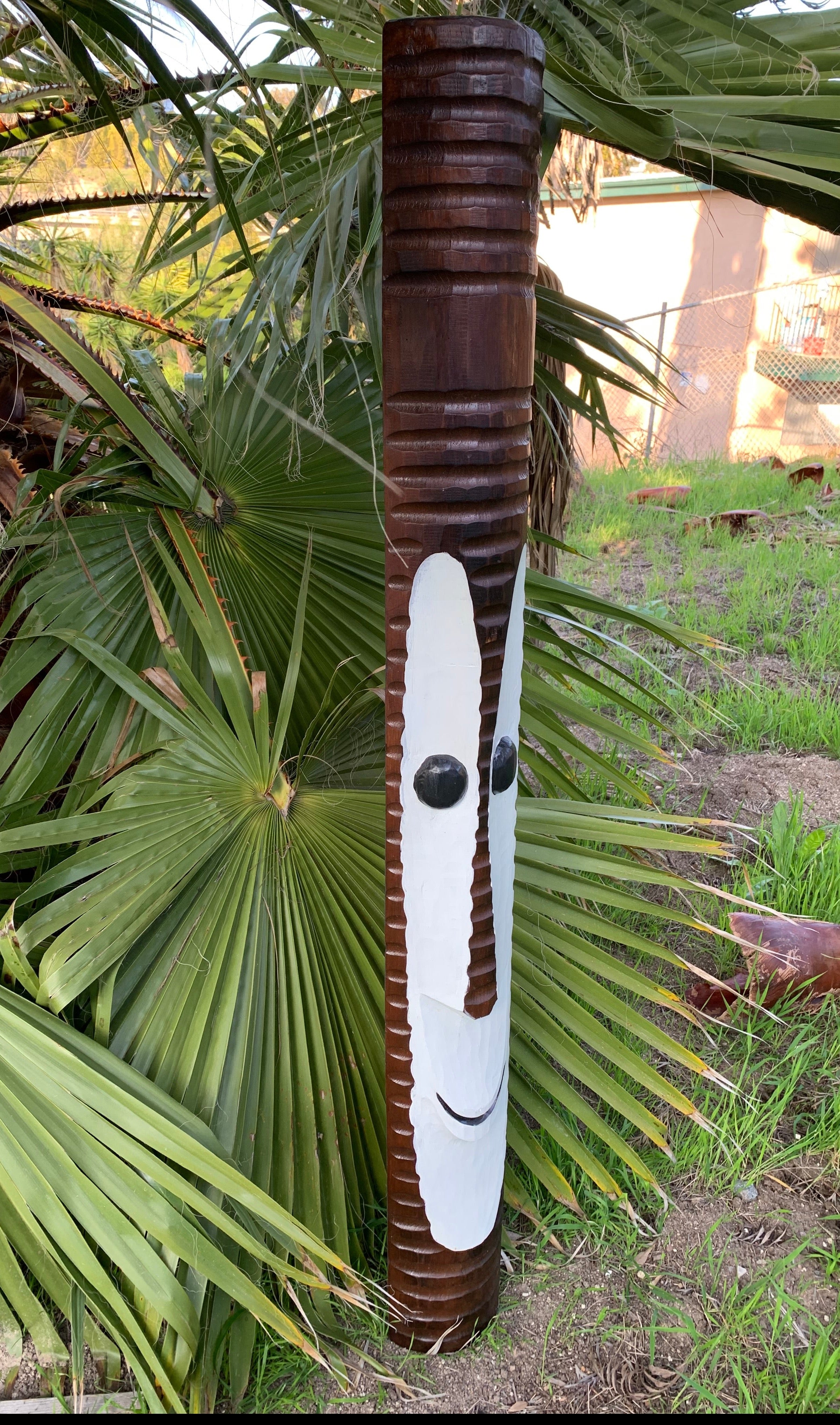 Carved wooden totem pole with a painted white face and black eyes among palm fronds