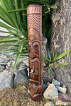 Tall carved wooden tiki totem with geometric patterns and a face in a garden setting