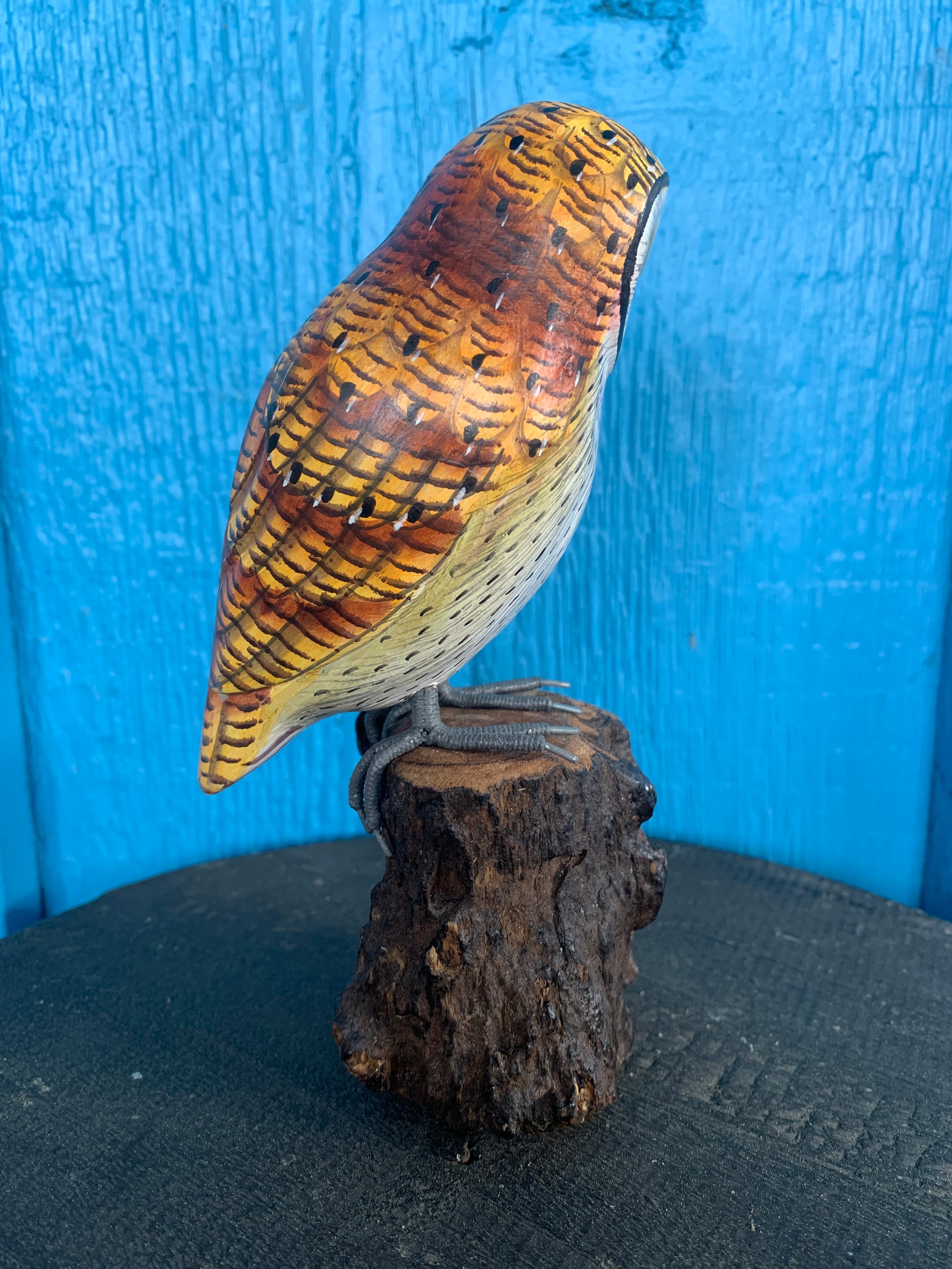 Carved wooden bird statue with orange-brown feathers perched on a rough log against a blue background.