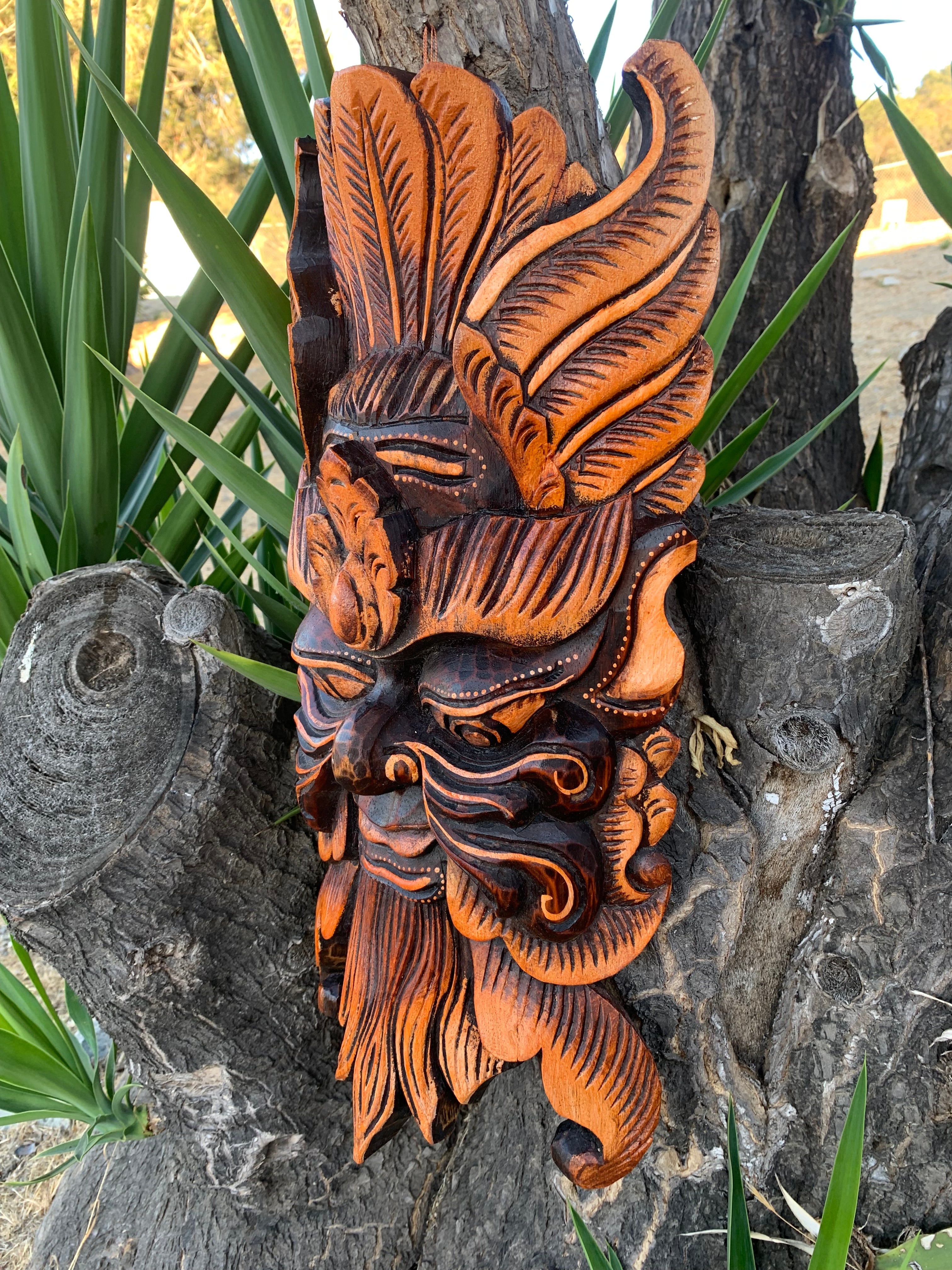 Orange and black carved wooden mask with feathered headdress mounted on a tree trunk