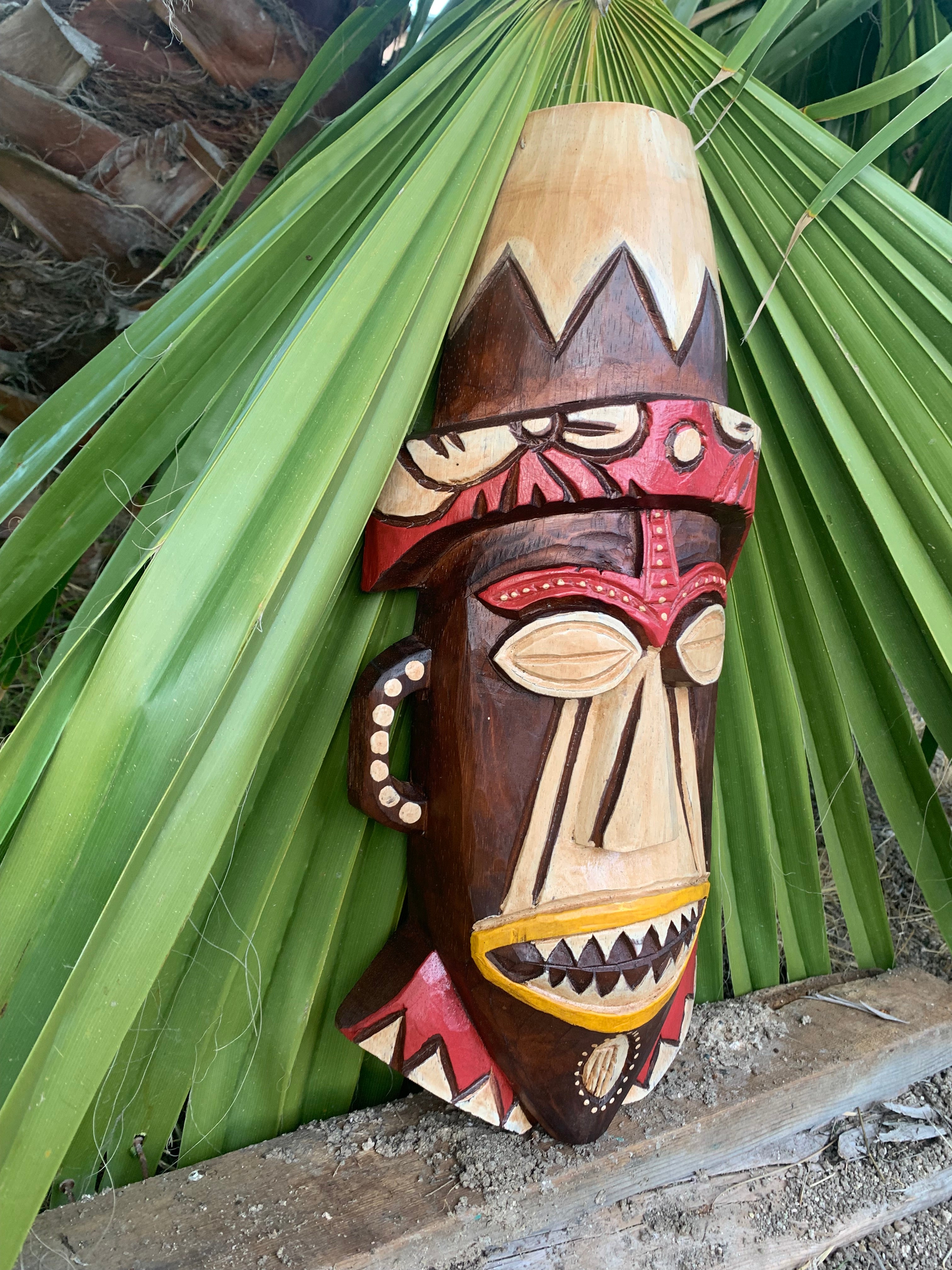 Colorful carved wooden tiki mask with geometric patterns resting against palm leaves