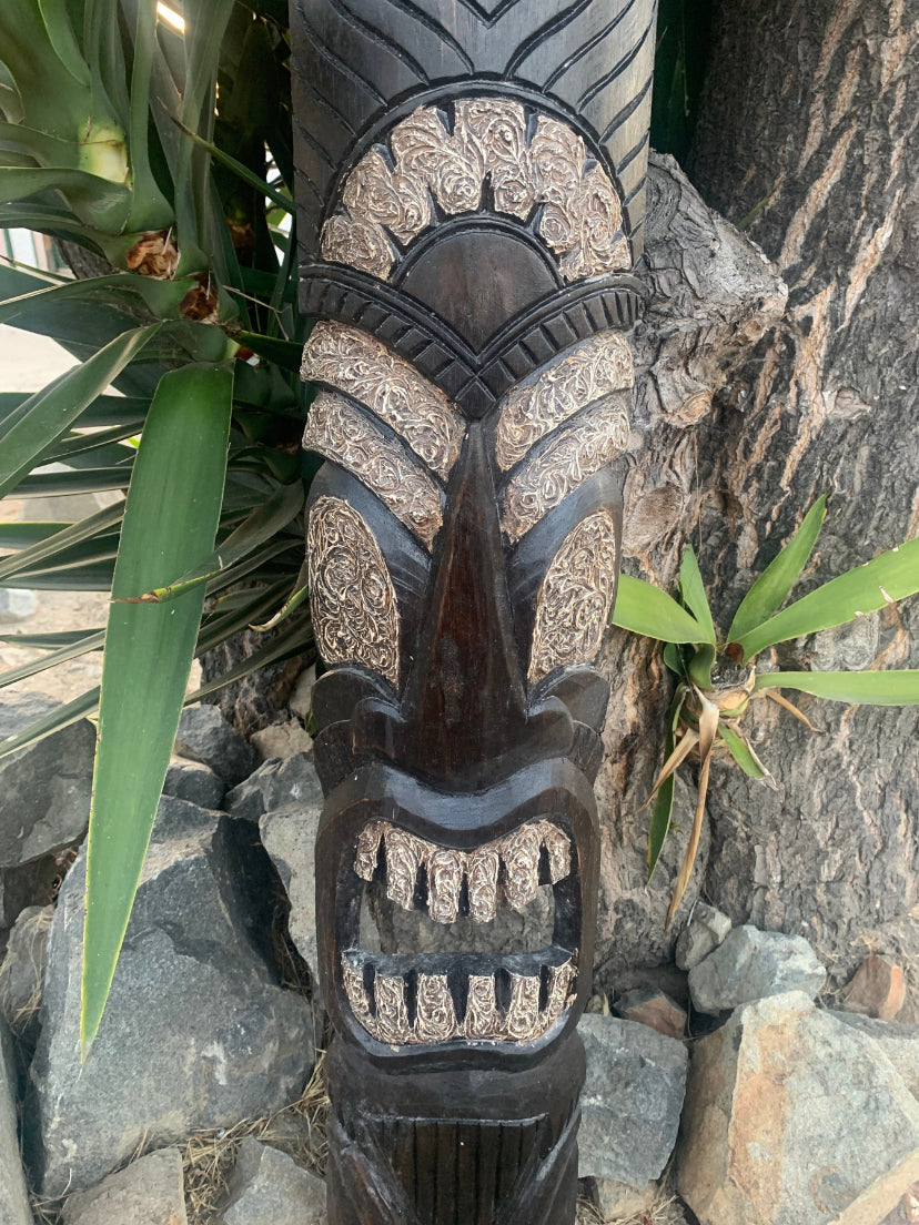 Carved wooden tiki mask with intricate patterned panels and a wide open mouth, outdoors.