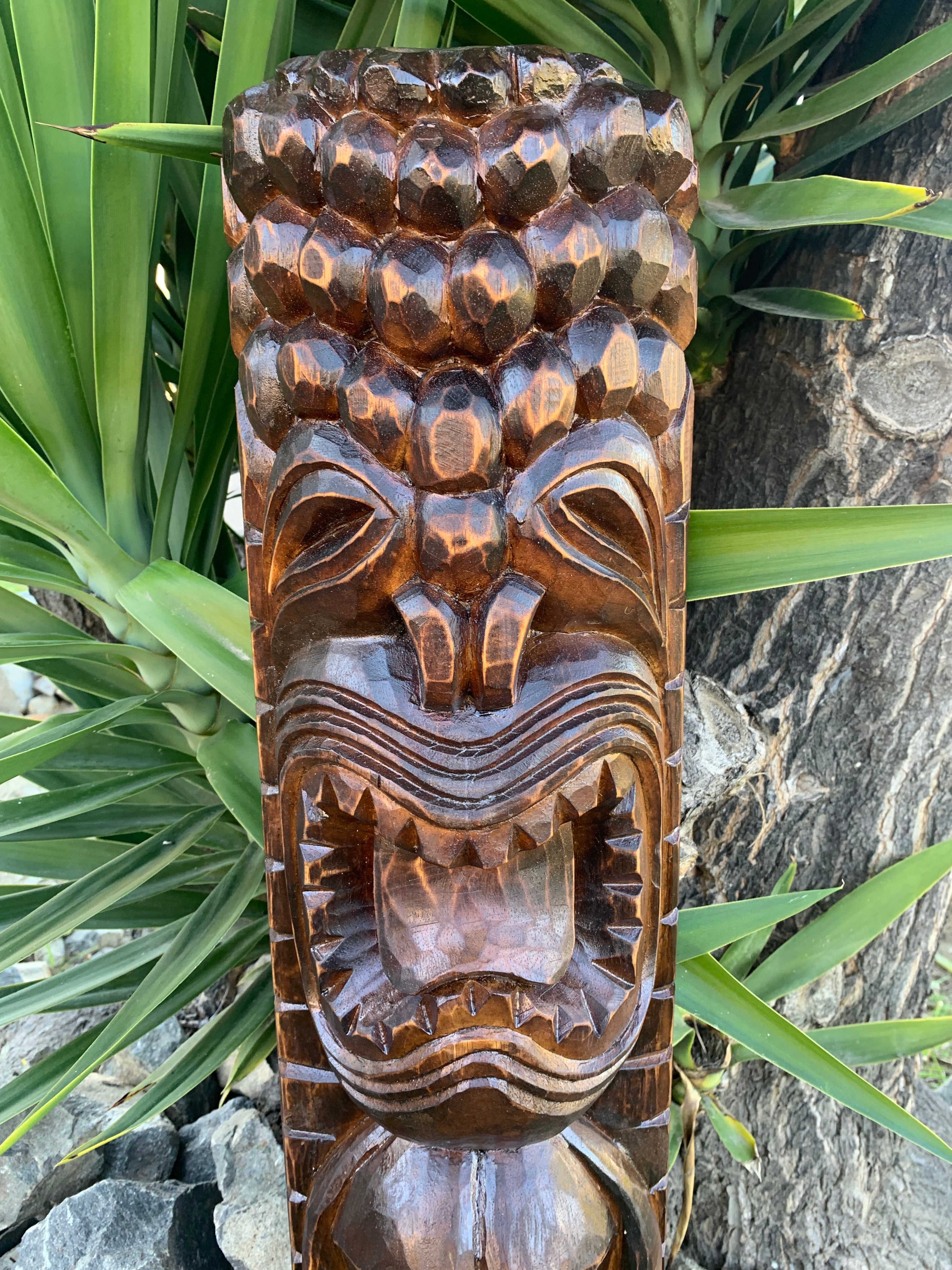 Carved wooden tiki mask with an open mouth and textured head among green leaves.