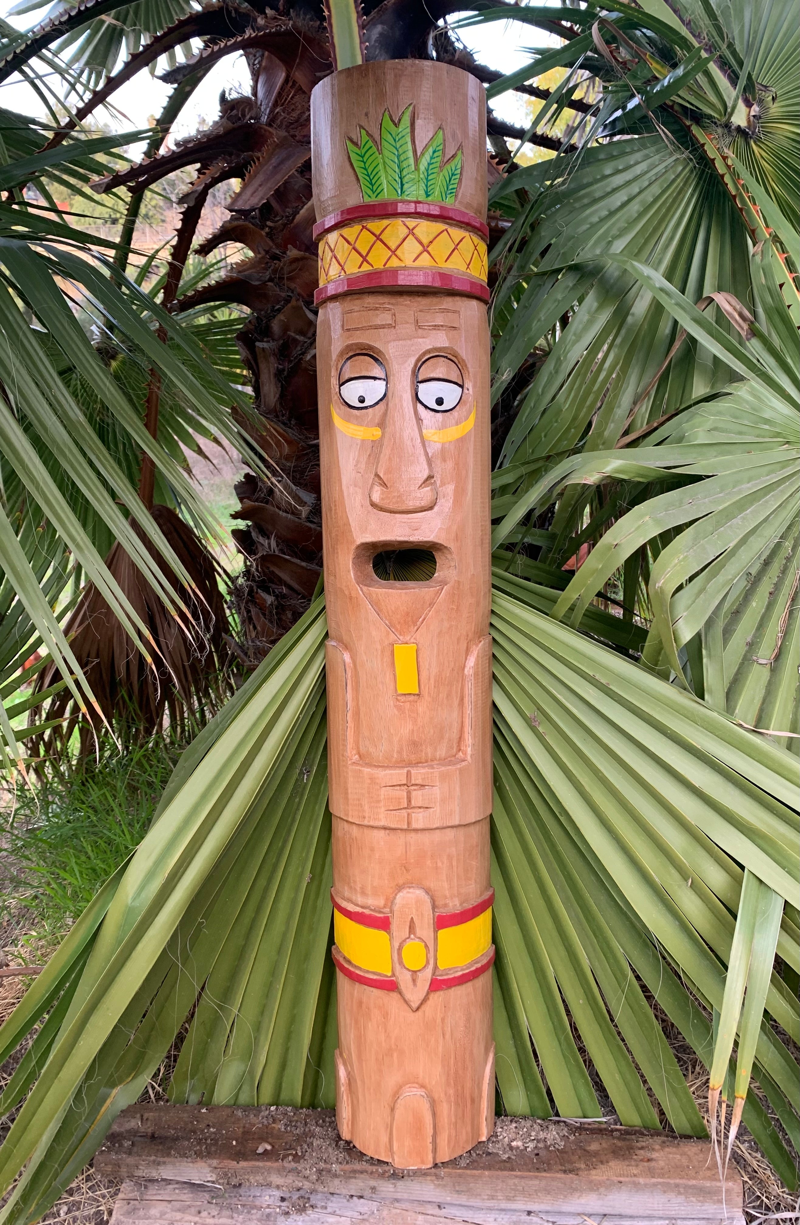 Carved wooden tiki statue with painted face and hat among palm leaves.