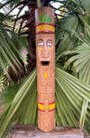 Carved wooden tiki statue with painted face and hat among palm leaves.