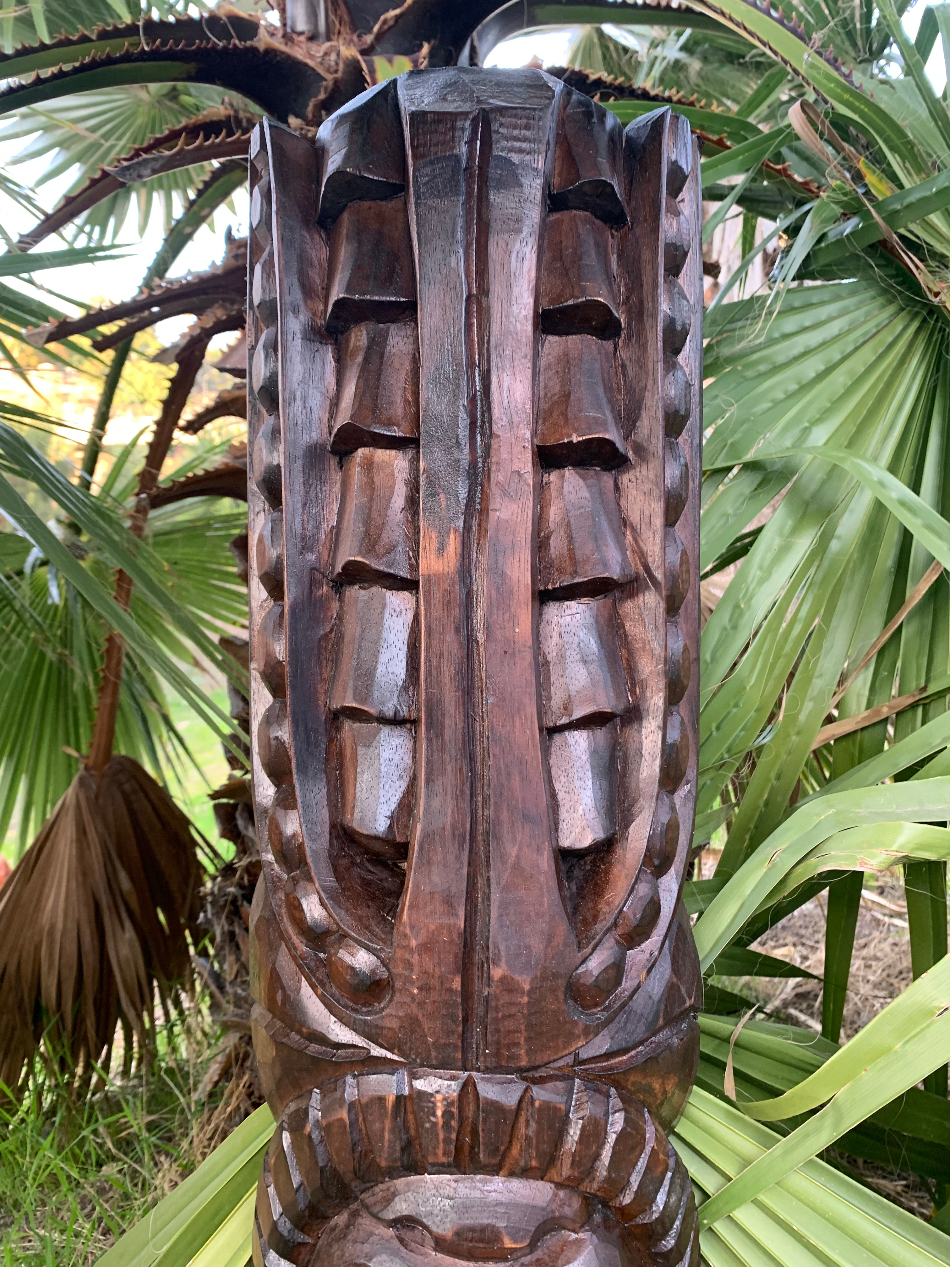 Close-up of a dark wooden tiki statue with geometric carvings in a tropical garden.