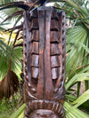 Close-up of a dark wooden tiki statue with geometric carvings in a tropical garden.