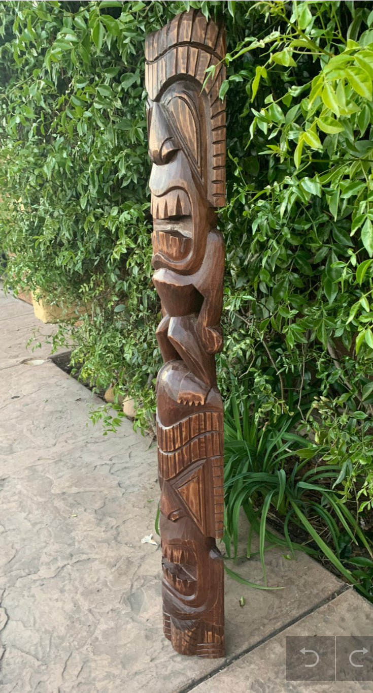 Carved wooden Tiki statue outdoors beside a green hedge.