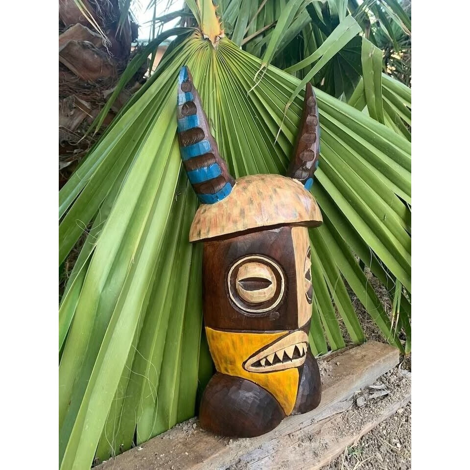 Carved wooden tiki statue with blue horn accents leaning against tropical palm leaves.