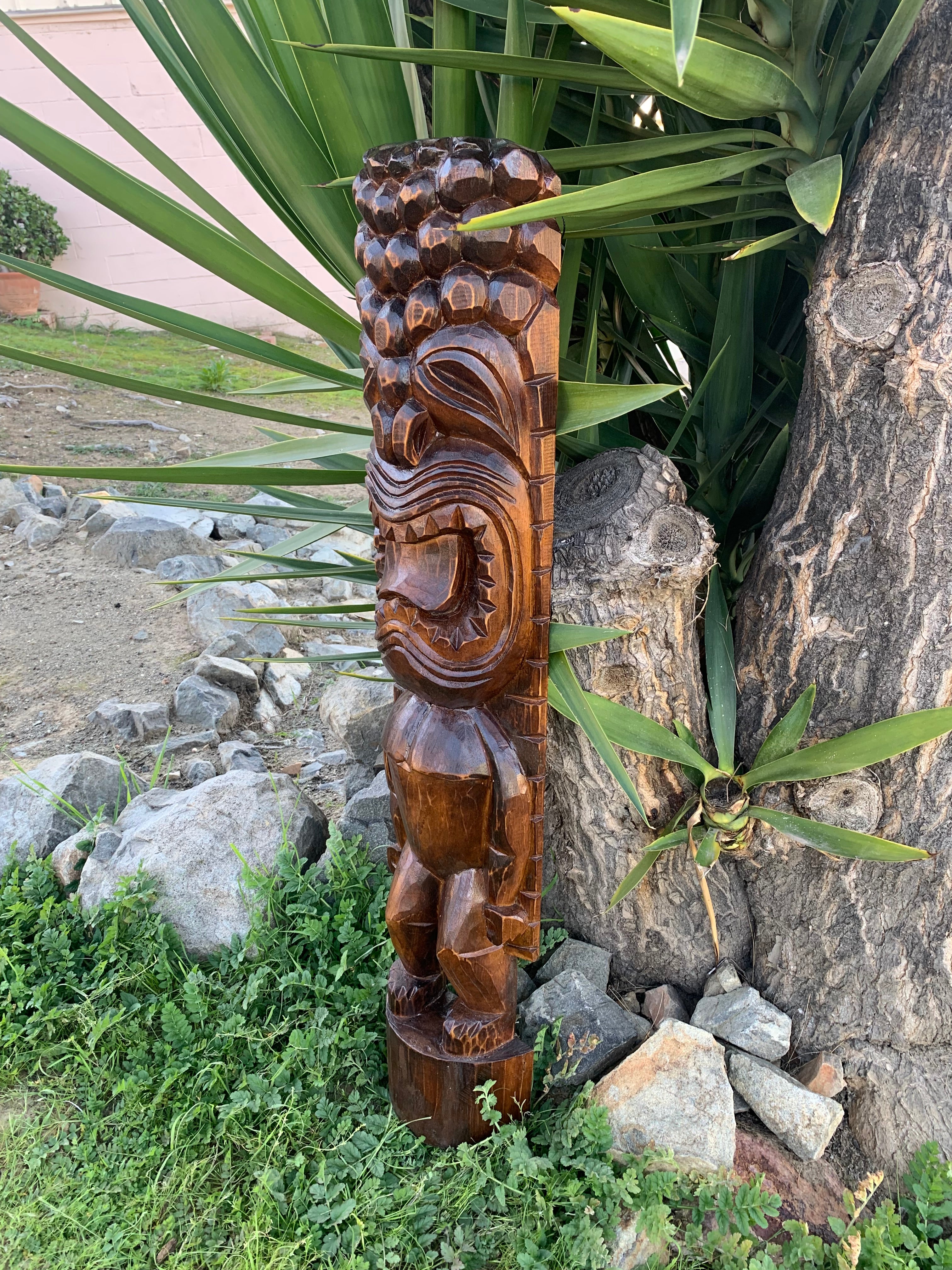 Hand-carved wooden tiki totem pole outdoors beside a tree in a garden.